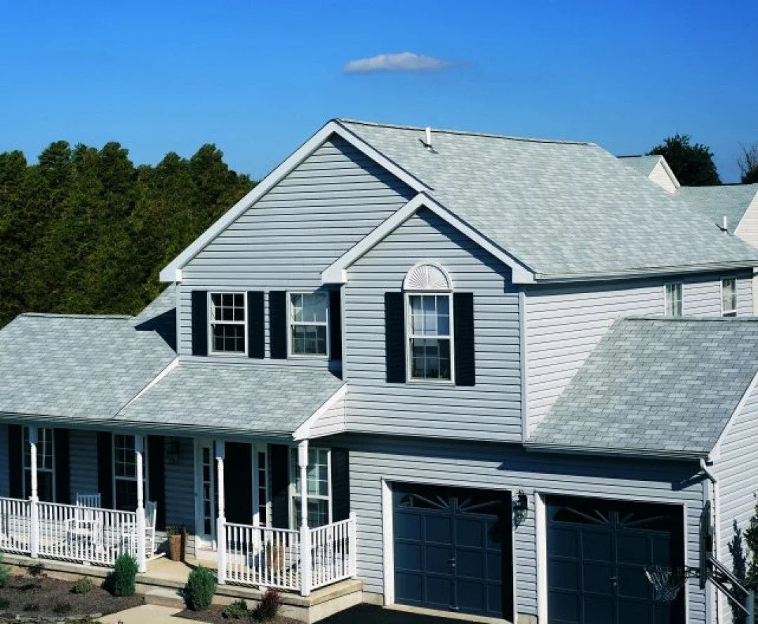 A two-story house with gray siding, black shutters, and a gray shingle roof, featuring a front porch with white railing, two garage doors, and surrounded by a well kept yard with small shrubs.