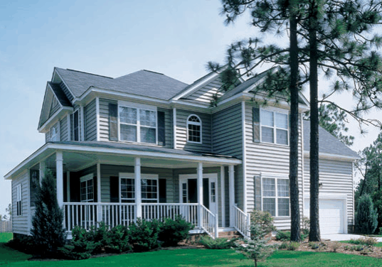 A two-story suburban house with vinyl siding, a front porch, and a two-car garage, surrounded by trees and a neatly maintained lawn.