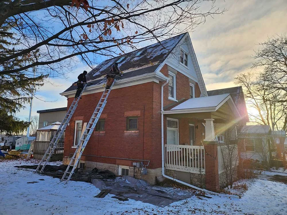 Two workers on ladders repairing the roof of a brick house during sunset on a snowy day.