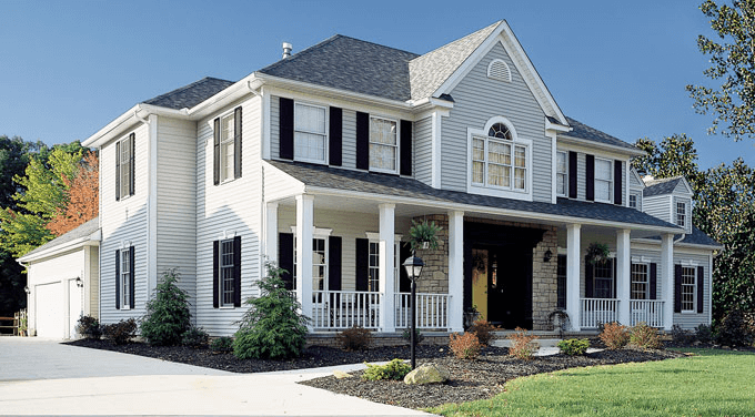 Large white two-story house with black shutters, front porch with railing, and a landscaped yard with trees and bushes.