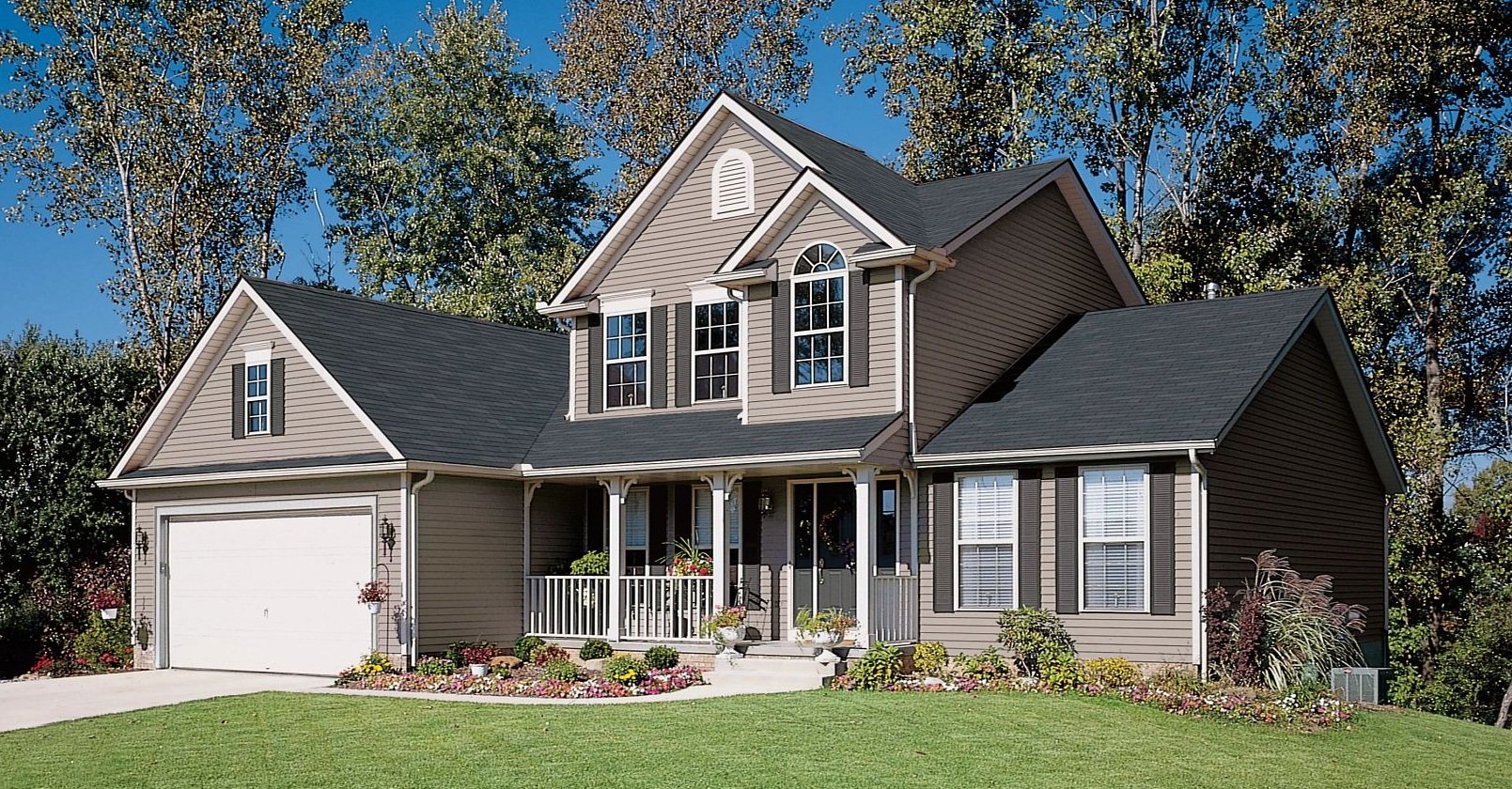 A two-story house with beige siding and black shutters, featuring a front porch with white railings, a white garage door, and a well-manicured lawn. There are multiple windows, including arched and rectangular ones, and trees with fall foliage in the background.