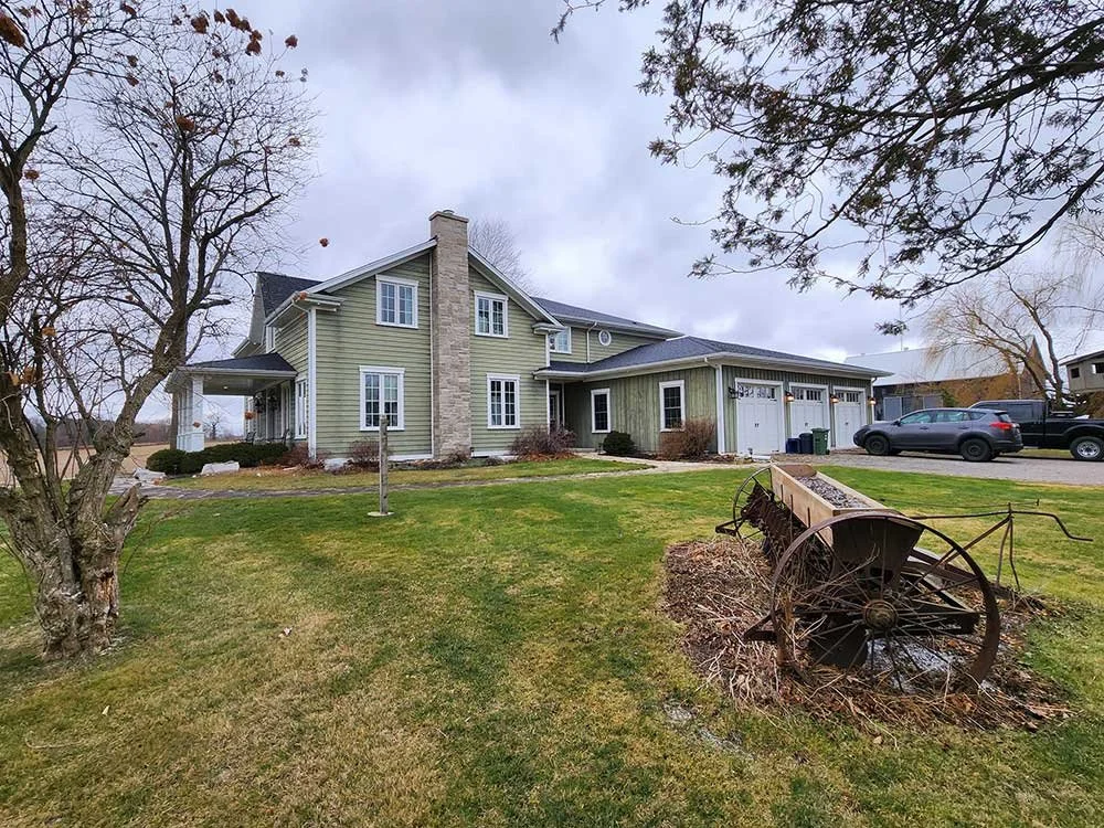 A two-story green house with white trim and attached garage, surrounded by a lawn with trees and a rusty old wagon.
