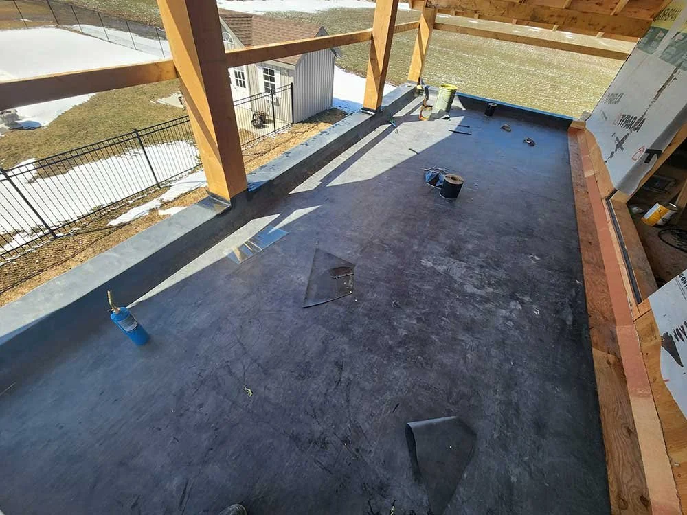 Construction site of a building balcony or deck with wooden framing and a concrete foundation, overlooking a yard with a fence and patchy snow.