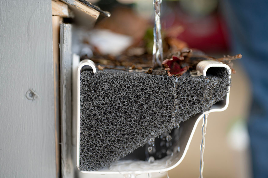 A close-up of a house gutter filled with leaves, dirt, and debris, with water spilling over the edge.