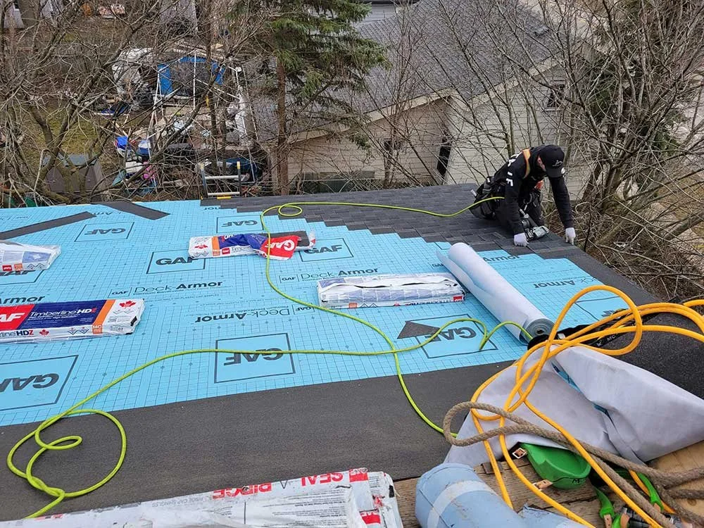 A roofer installing underlayment on a residential roof, surrounded by construction tools and materials, with trees and neighboring houses in the background.