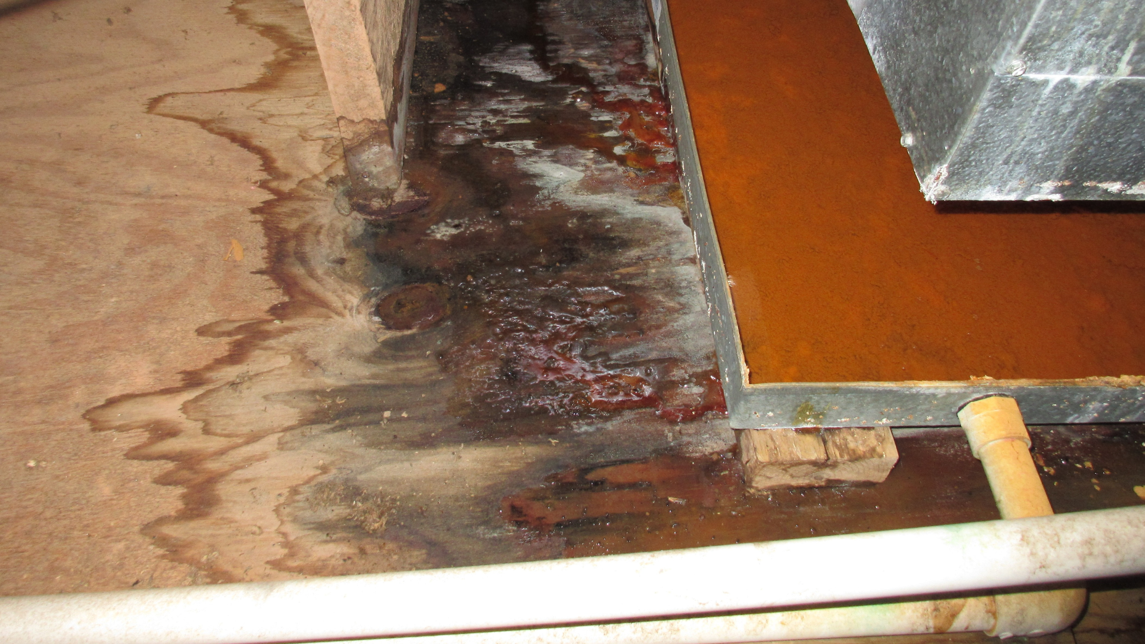 Close-up of a metal stain and water damage on a wooden floor with a small brick beneath a metal structure.