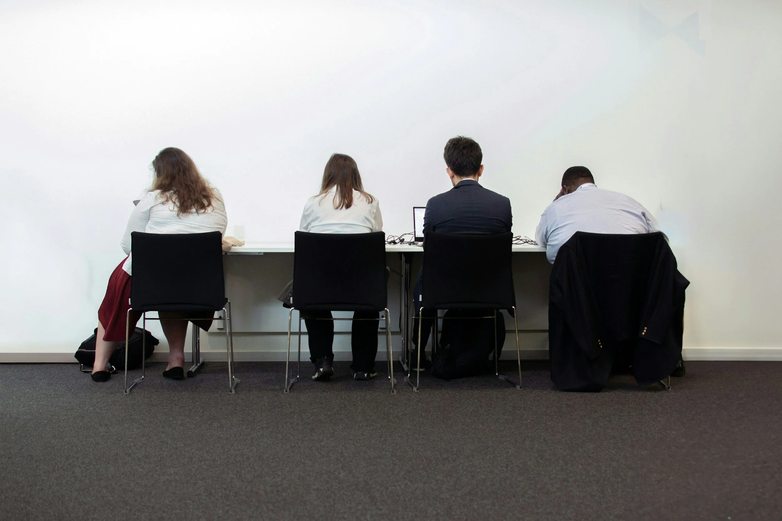 Four individuals sitting at a table in front of a blank white wall, viewed from behind. Two women on the left and two men on the right, two with laptops open in front of them.