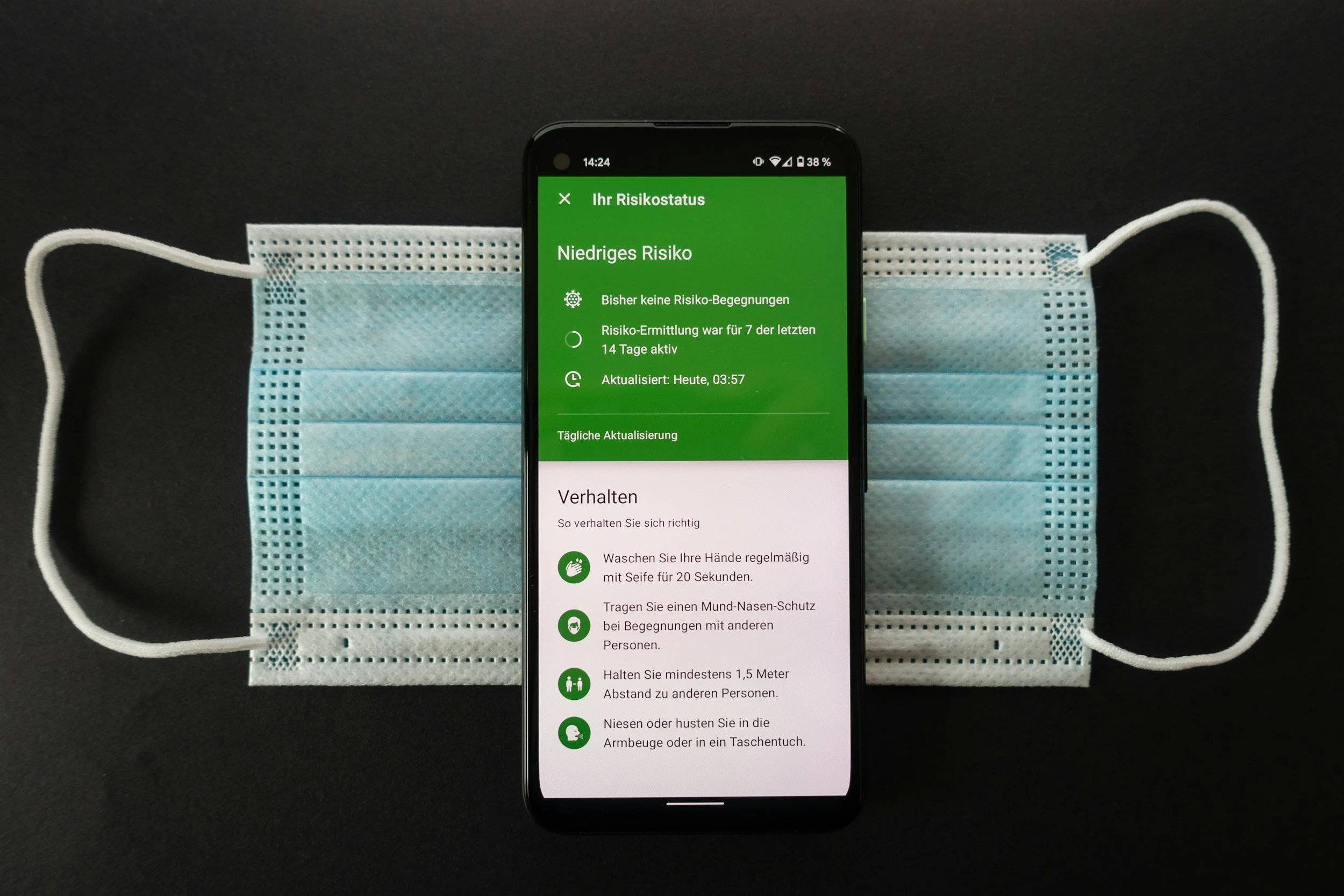 Smartphone placed on a black surface with a disposable face mask behind it. The smartphone screen displays a health app with information about COVID-19 risk status and safety guidelines in German.