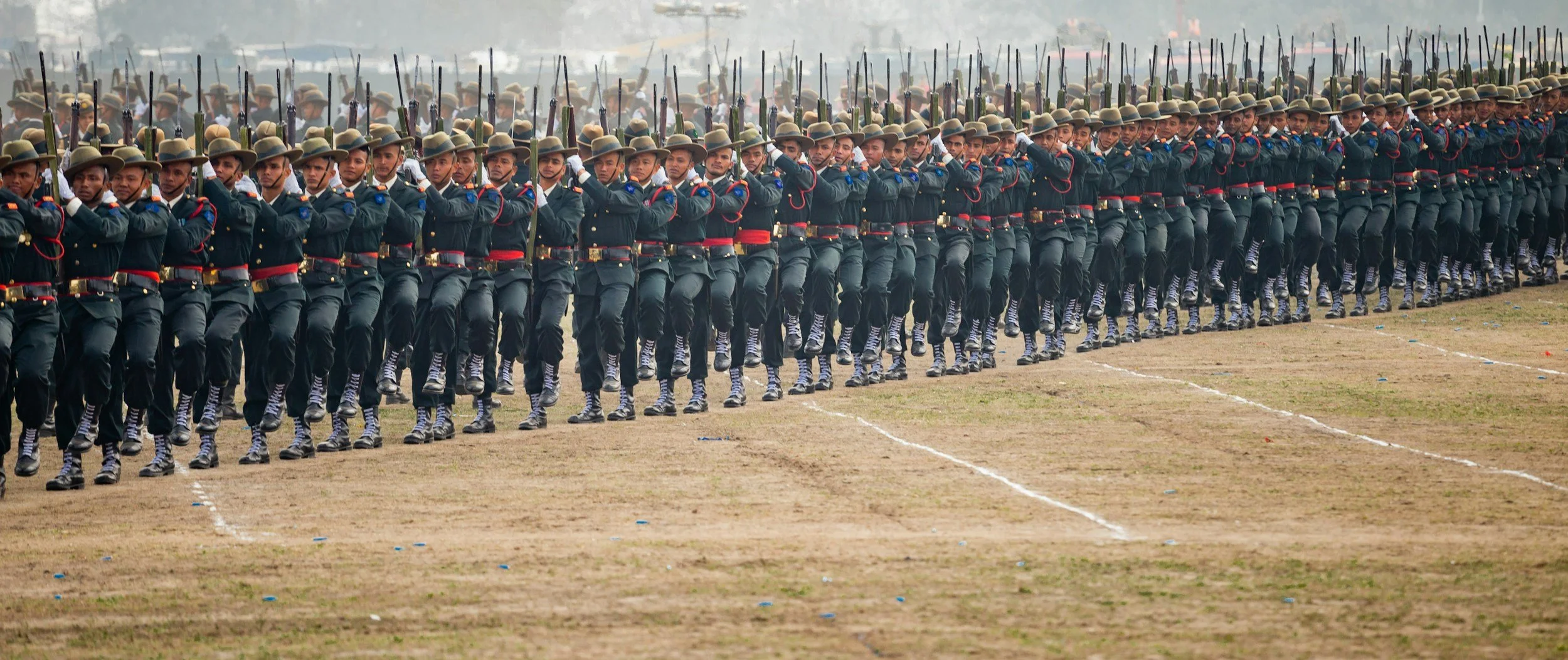 A large group of soldiers in uniform marching in formation during a parade, with rifles on their shoulders and a dusty field beneath them.