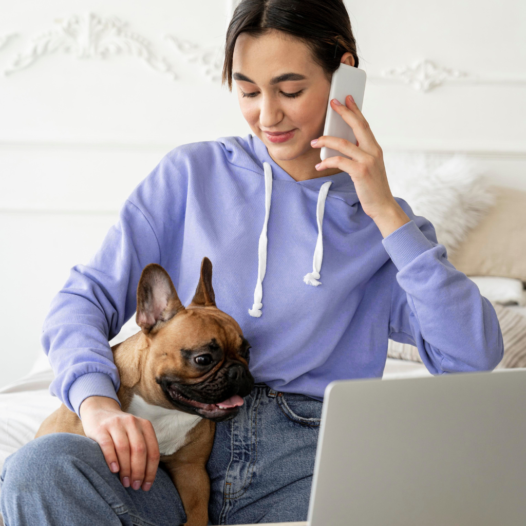 A young woman in a lavender hoodie sitting on a bed, holding a French bulldog on her lap, talking on a cell phone, with a laptop in front of her.