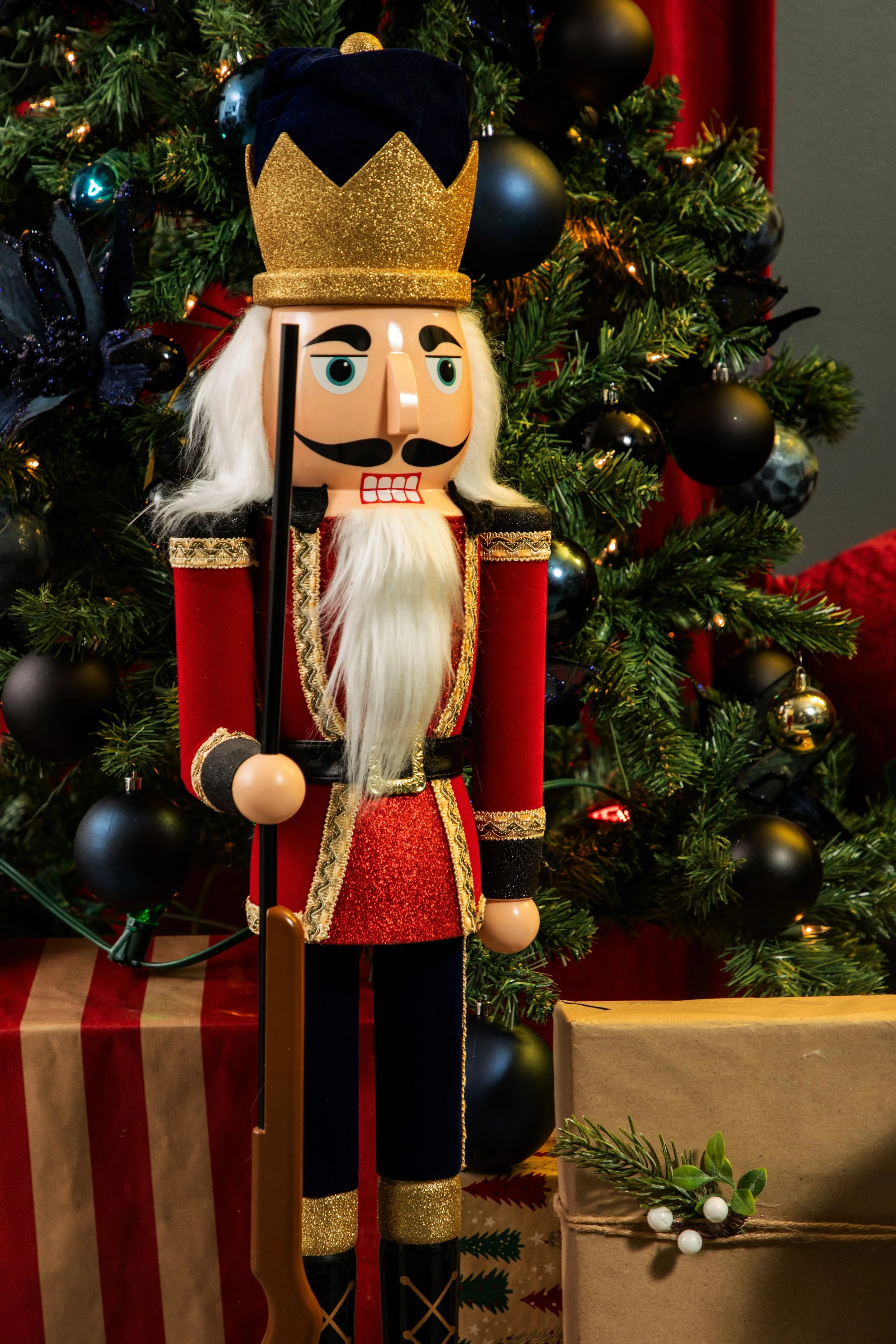 Un soldat en bois décoratif de Noël avec un uniforme rouge et or, une barbe blanche, un chapeau avec une touche de noir et une couronne dorée, tenant une lance, devant un sapin de Noël décoré de boules noires et de guirlandes lumineuses, avec des cad