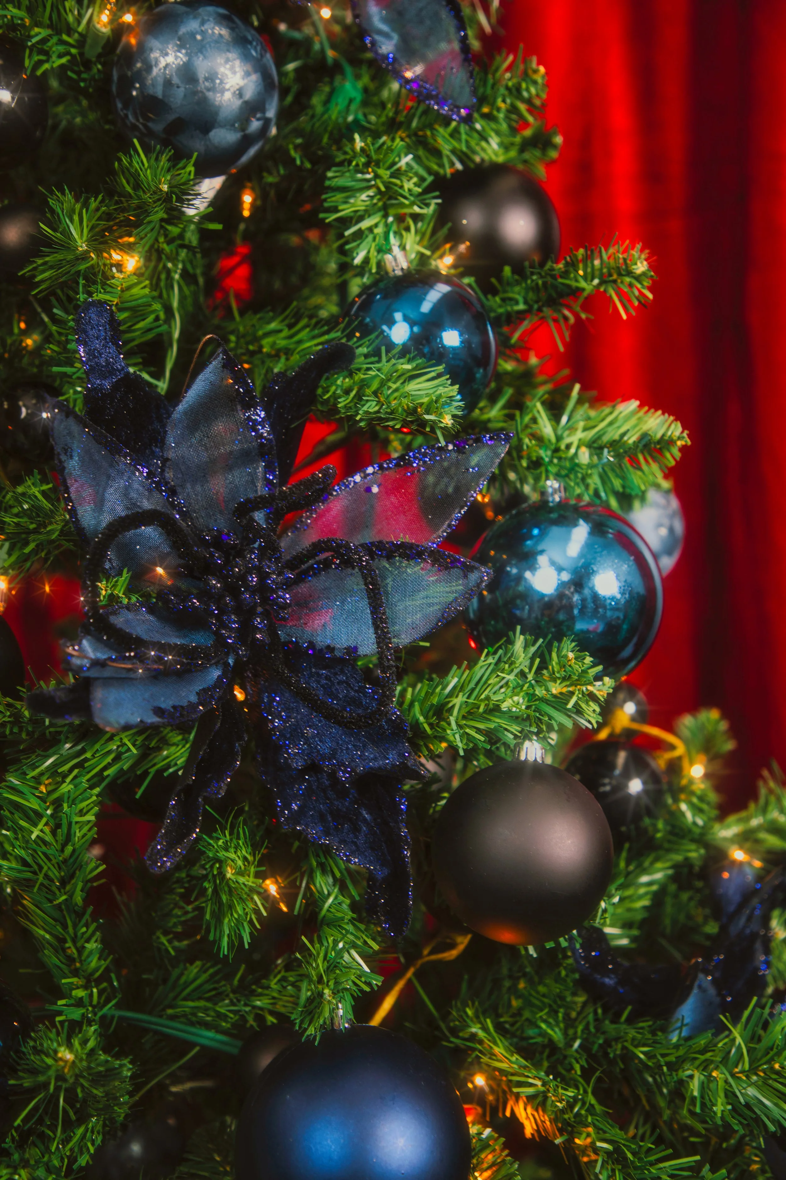 Décor de Noël avec une branche de sapin ornée de boules colorées, une grande fleur décorative noire et un fond de rideau rouge.