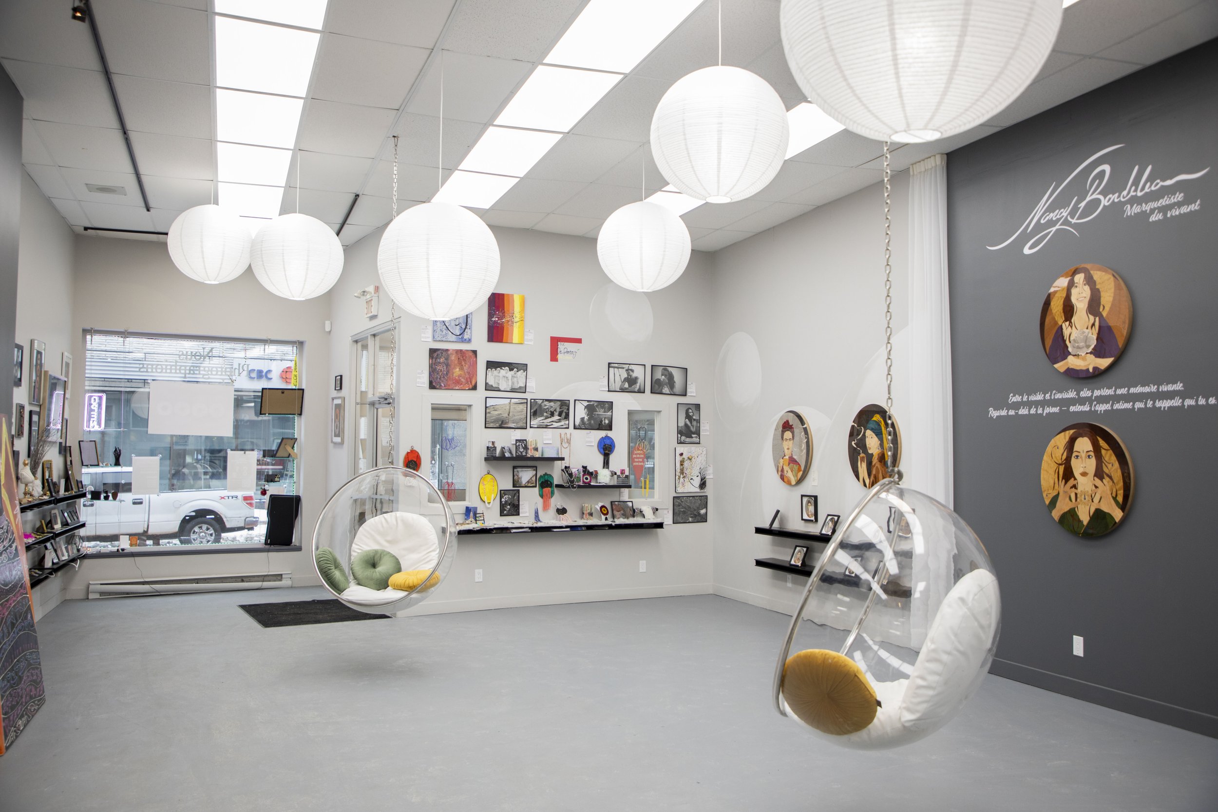 Une galerie d'art avec des œuvres sur les murs, deux bancs suspendus en forme de bulles transparentes avec coussins colorés, et des lampes rondes suspendues au plafond.