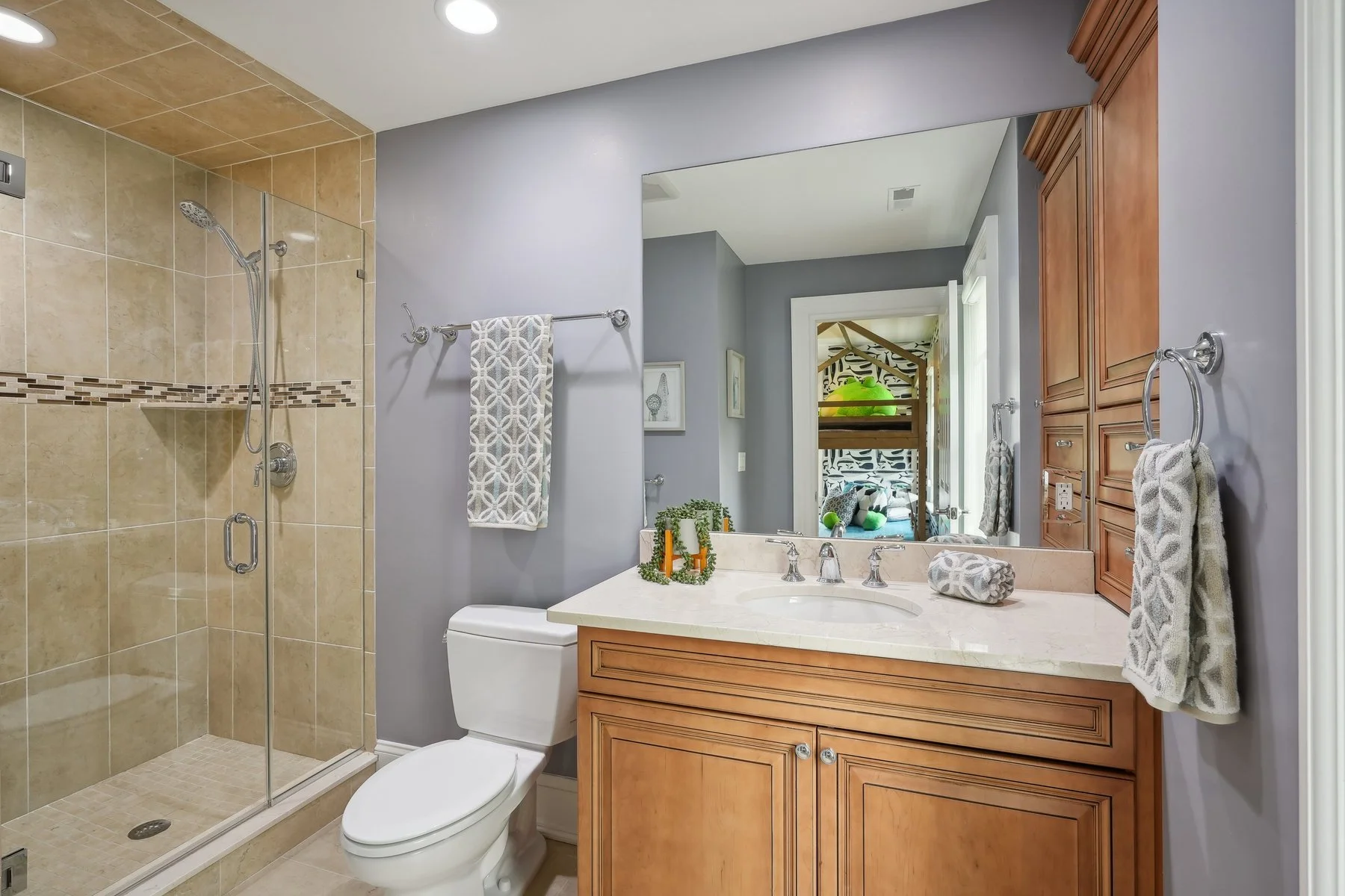 Bathroom with a glass shower enclosure, a white toilet, a wooden vanity with a marble countertop, a large mirror, and a view into a bedroom with bunk beds.