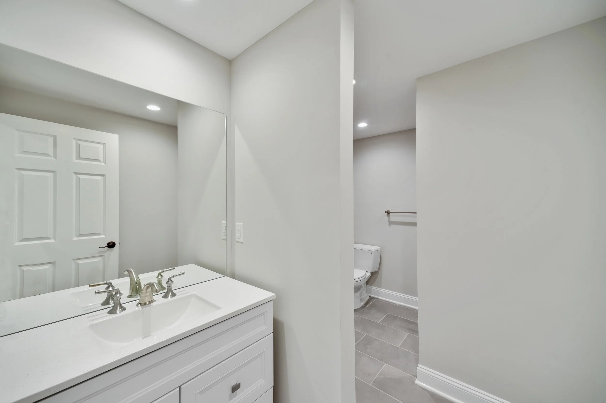 Bathroom with white vanity, faucet, large mirror, toilet, gray tile floor, white walls, recessed lighting, towel bar.