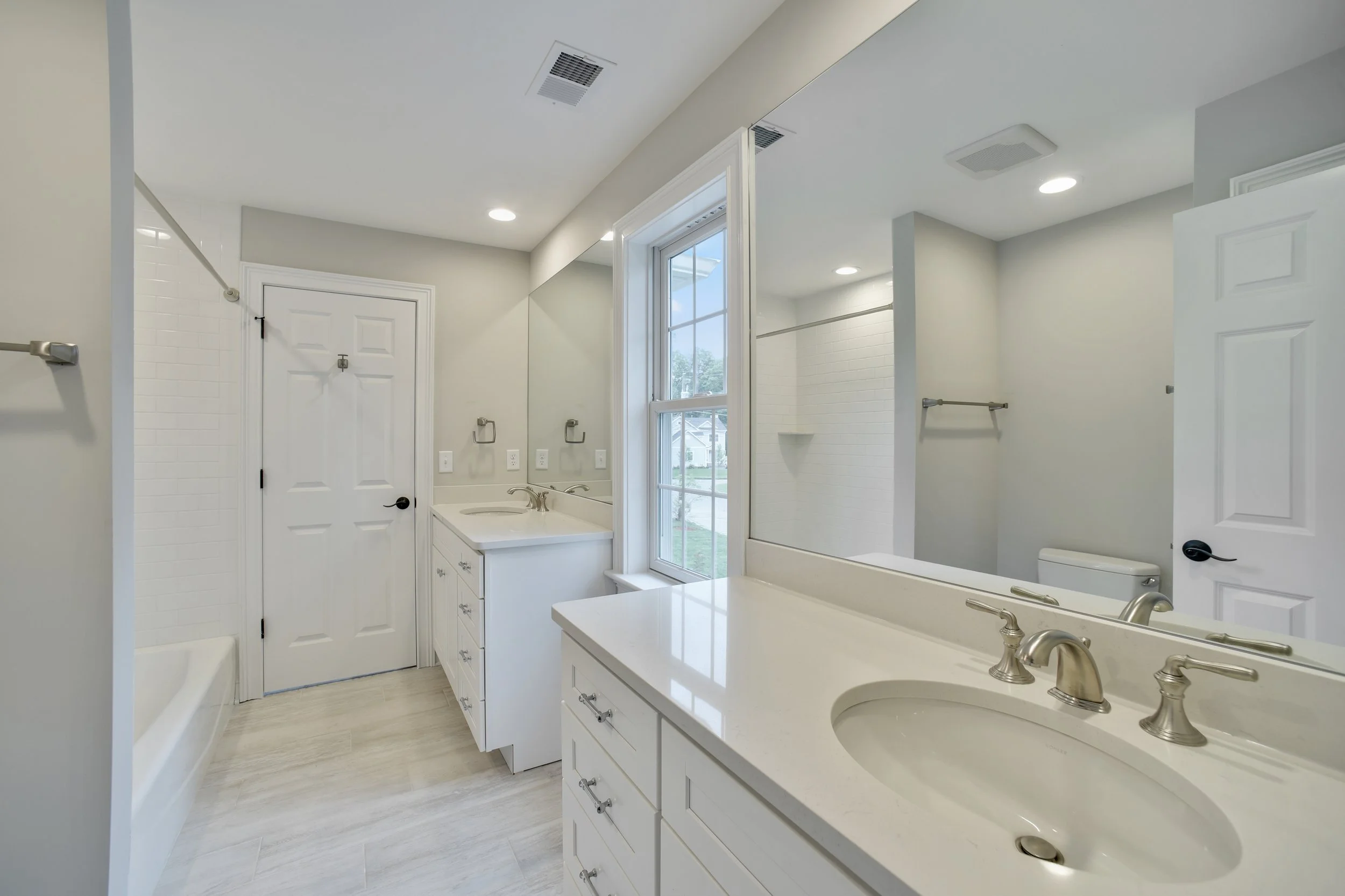 Interior view of a clean, modern bathroom with white cabinets, a large mirror, a vanity with a sink, a window, and a shower area.