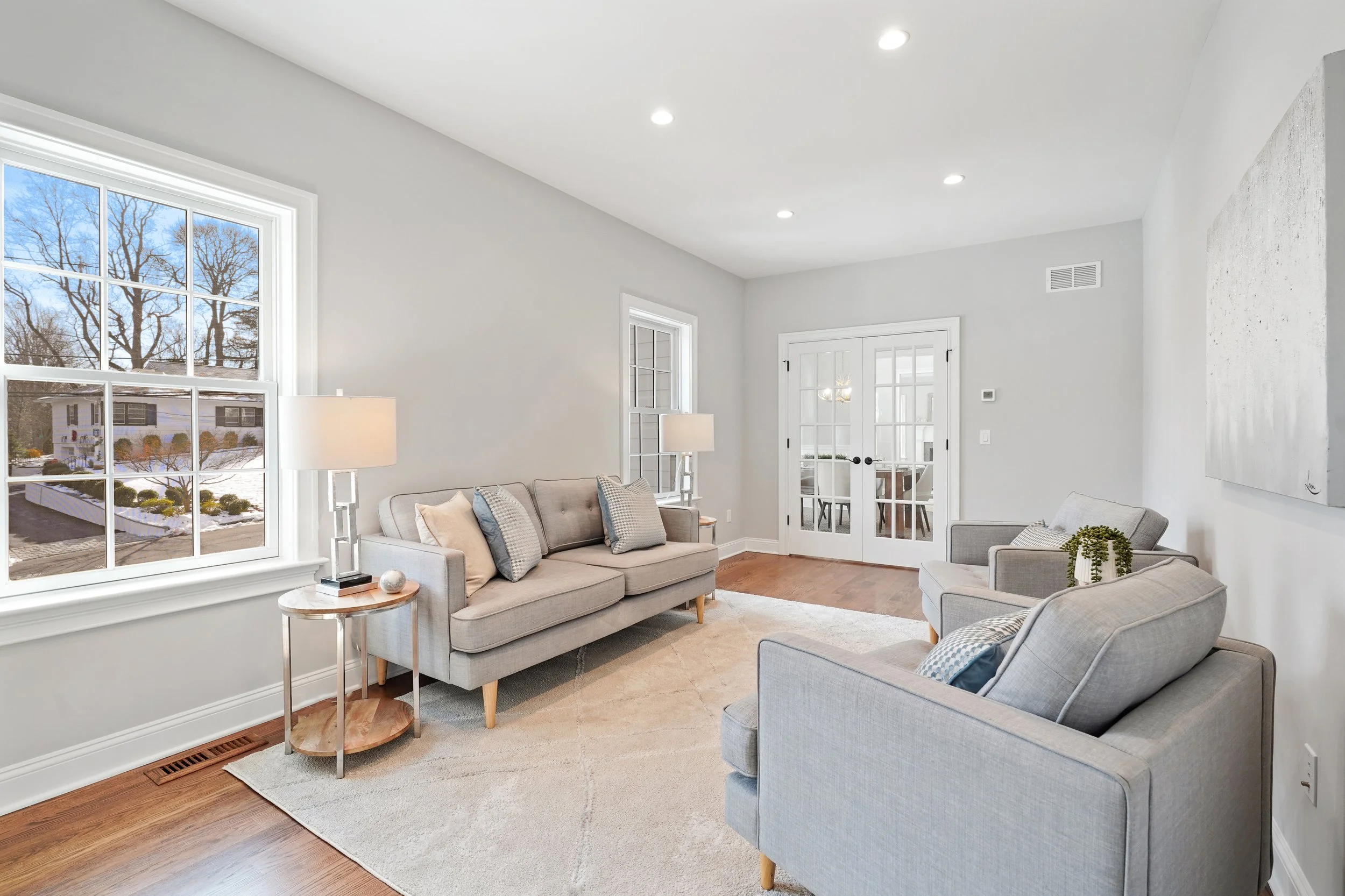 Living room with light gray sofa, two gray armchairs, white walls, hardwood floor, large window showing trees and snow, sliding glass door, side table with lamp, decorative pillows, and artwork on wall.