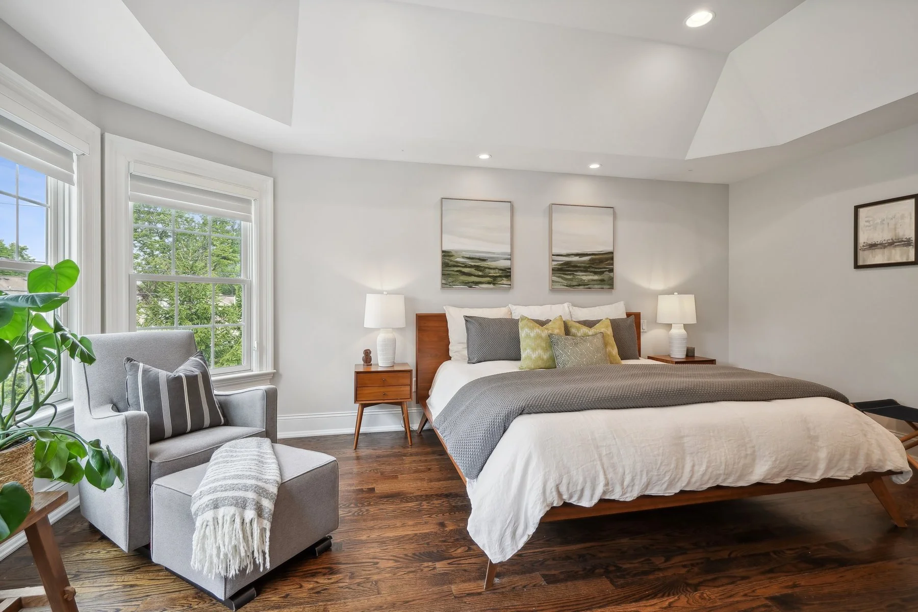 A modern bedroom with a large bed, nightstands with lamps, a gray armchair with a pillow and blanket, windows with lush greenery outside, and hardwood floors.