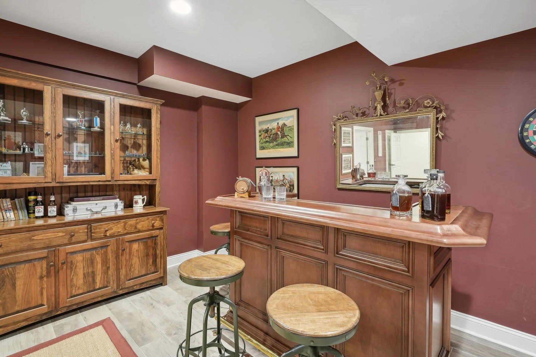 Home bar area with wooden cabinet, marble-topped bar counter, bar stools, framed pictures, and decorative bottle glasses on a maroon wall.