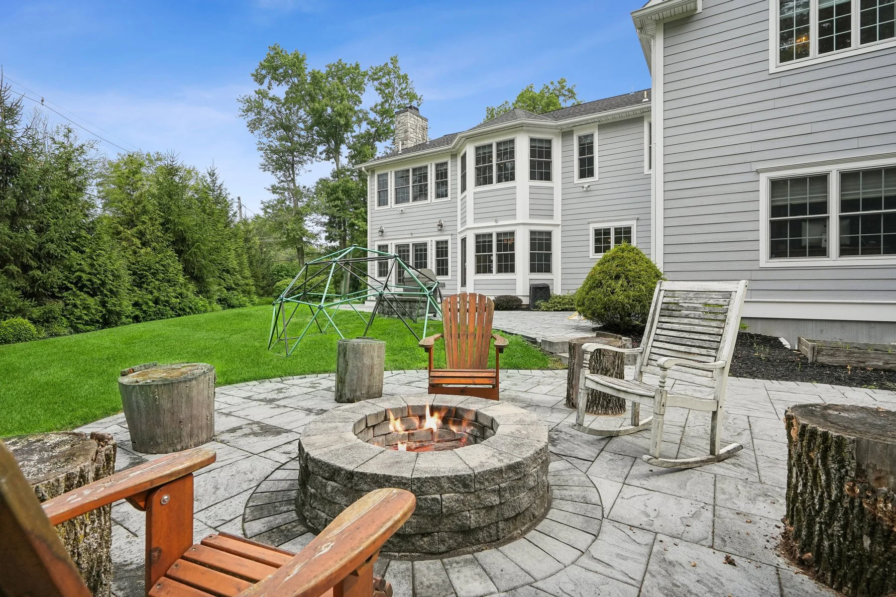 A backyard patio with a fire pit surrounded by wooden chairs and tree stump seats, with a grassy yard, a children’s climbing dome, and a light gray house with multiple windows in the background.