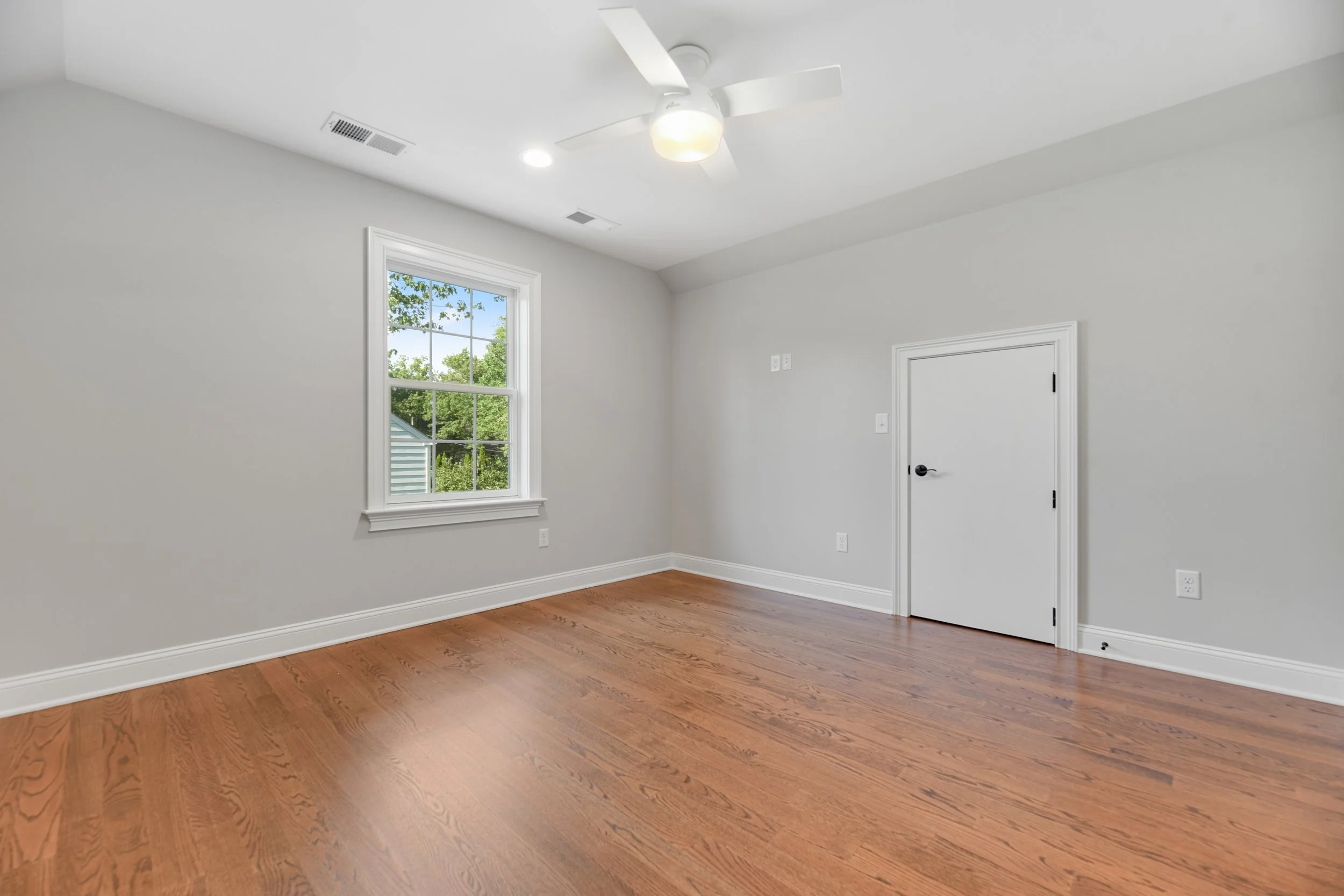 Empty room with hardwood floor, white walls, a window showing greenery outside, a ceiling fan, and a small door.