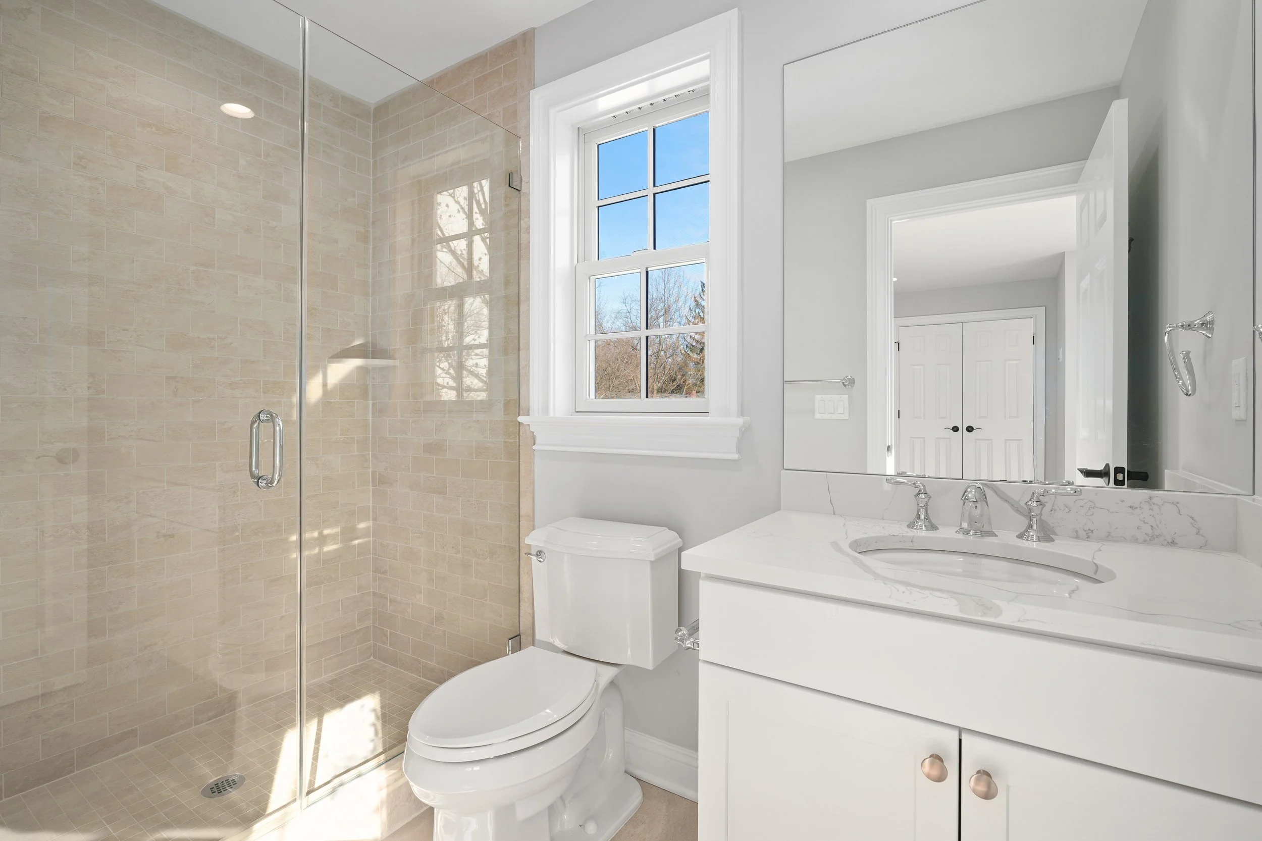 Bathroom with beige-tiled shower, white toilet, white vanity with marble countertop, large mirror, window overlooking trees