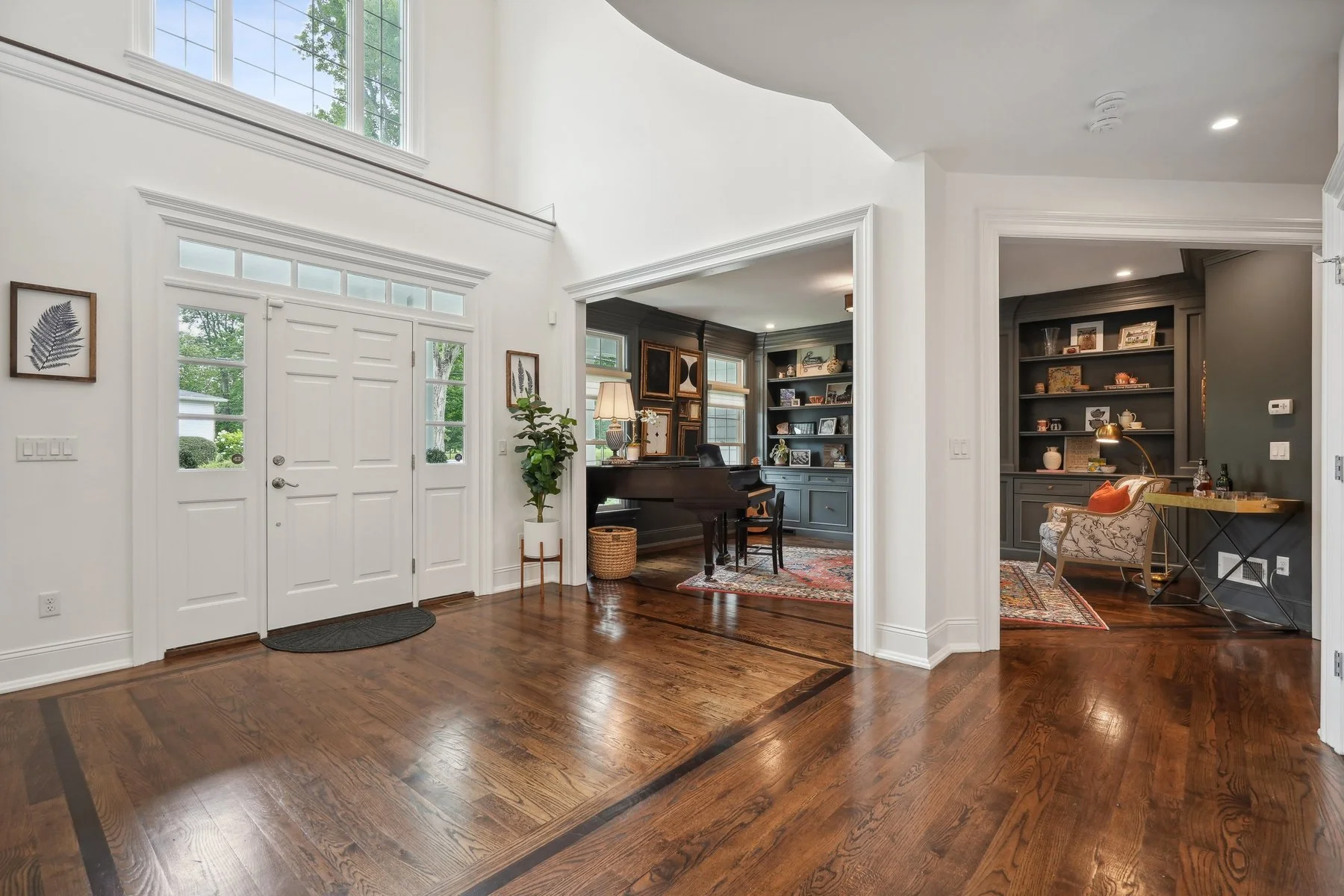 A spacious entryway and two connected rooms with hardwood floors, white walls, and large windows. The entry has a white front door with glass panels and a small round rug. The middle room features a dark-colored desk, shelves with decorative items, a