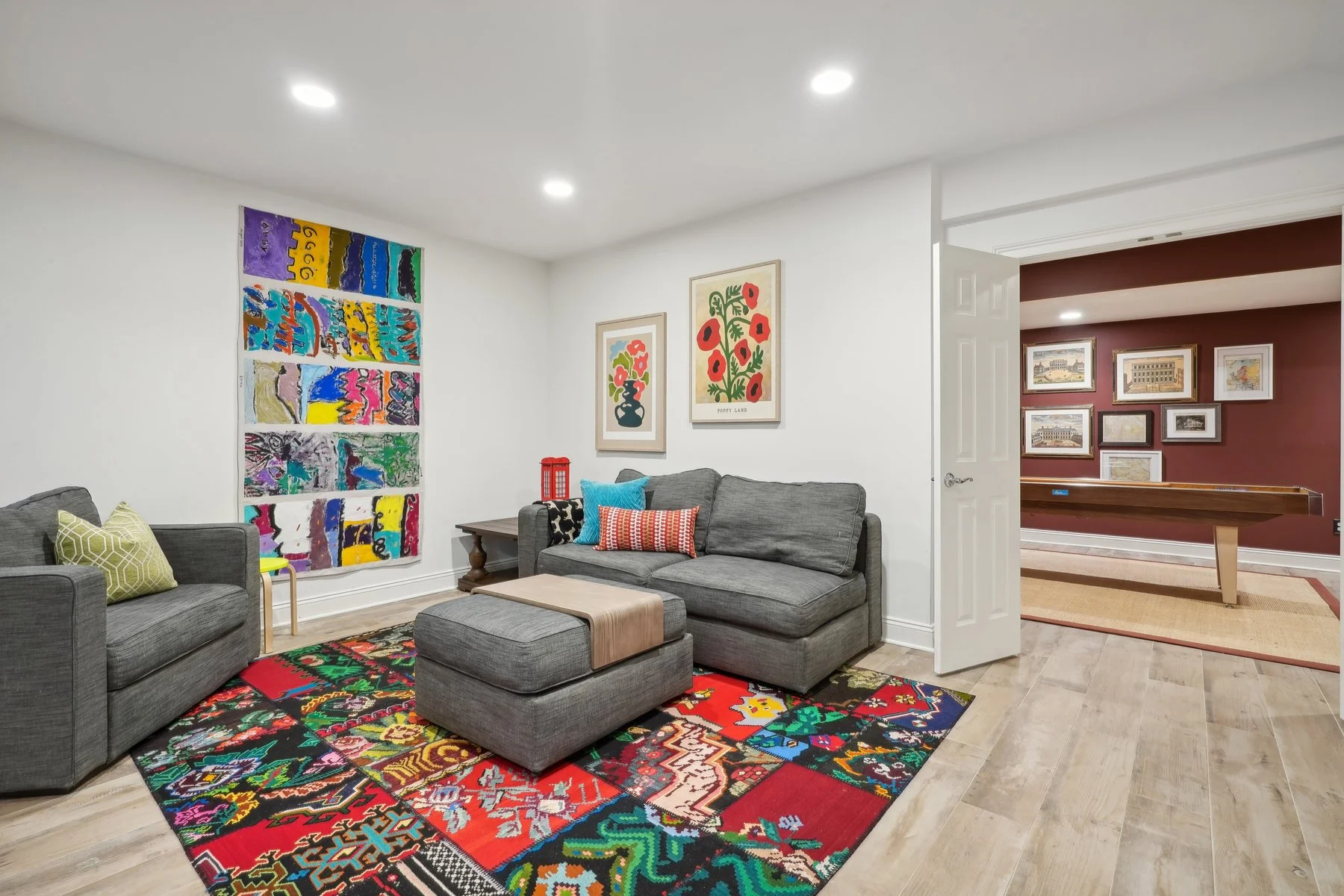 Living room with gray sofas, colorful artwork on the walls, a vibrant patterned rug, and a view into a game room with framed pictures and a shuffleboard table.