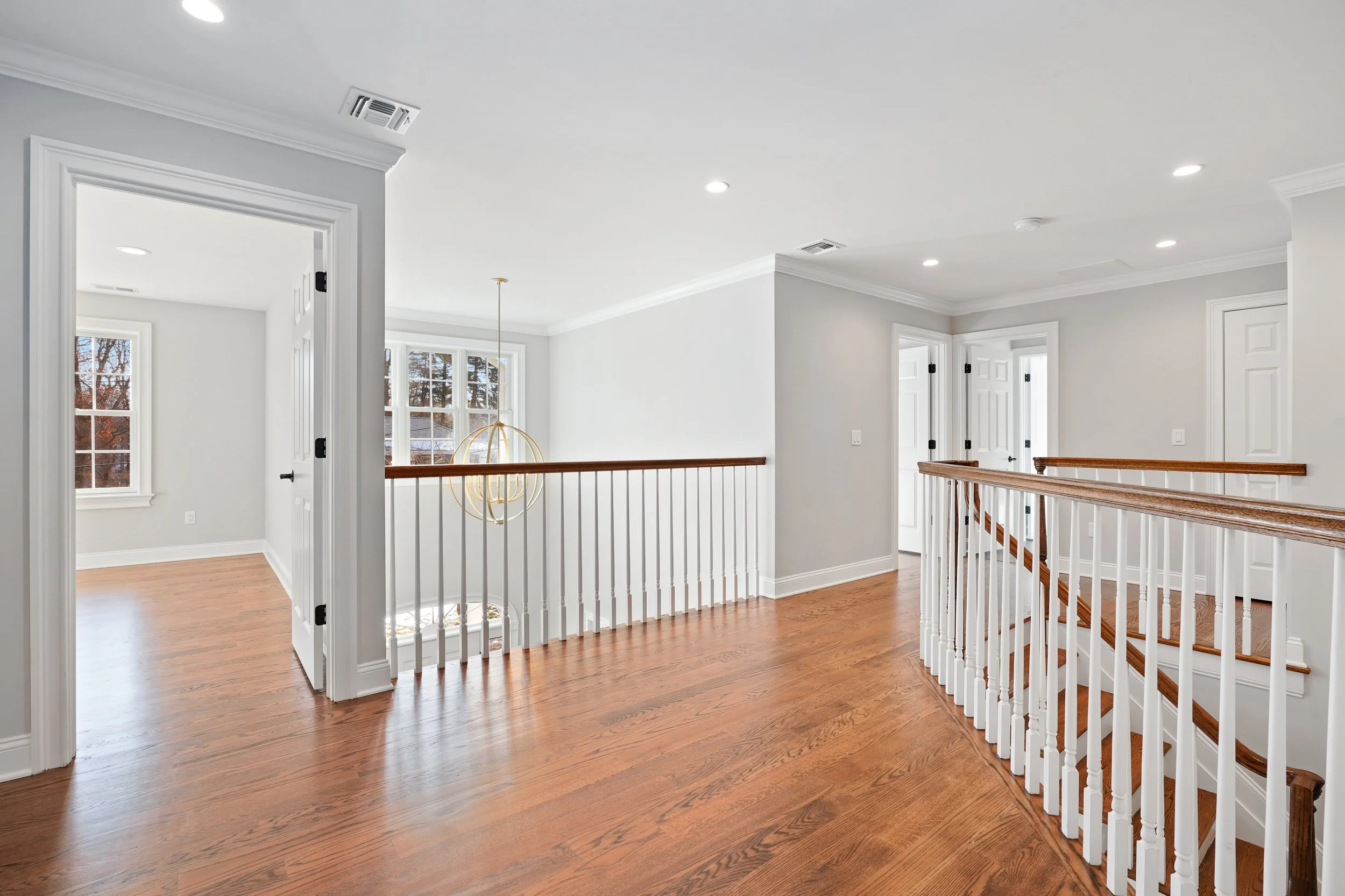 Bright interior of a modern home with hardwood floors, white walls, a staircase with wooden handrails, and multiple windows letting in natural light.