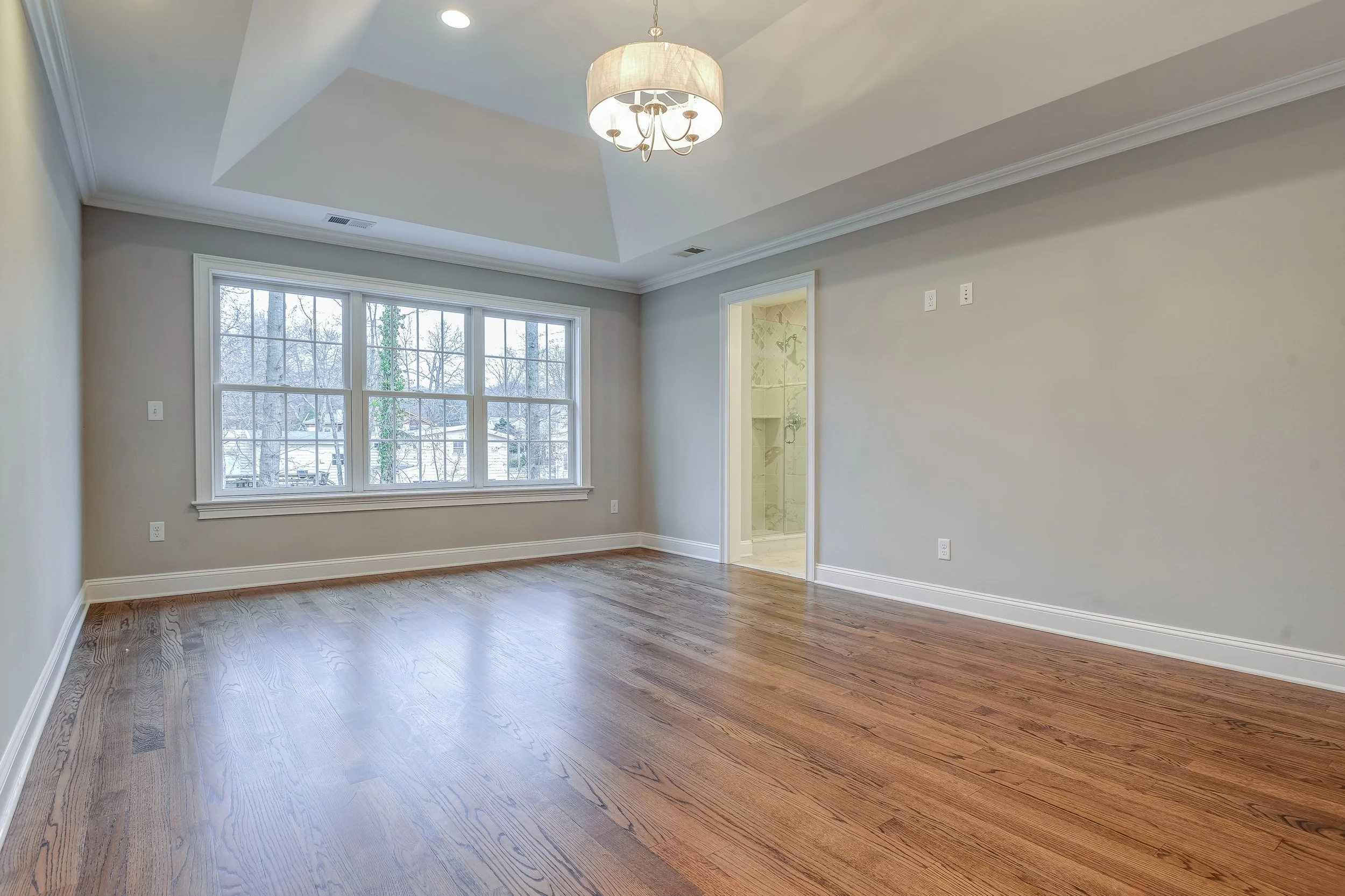 Empty living room with large window, hardwood floors, and an open bathroom door to a tiled shower, with a ceiling light fixture.