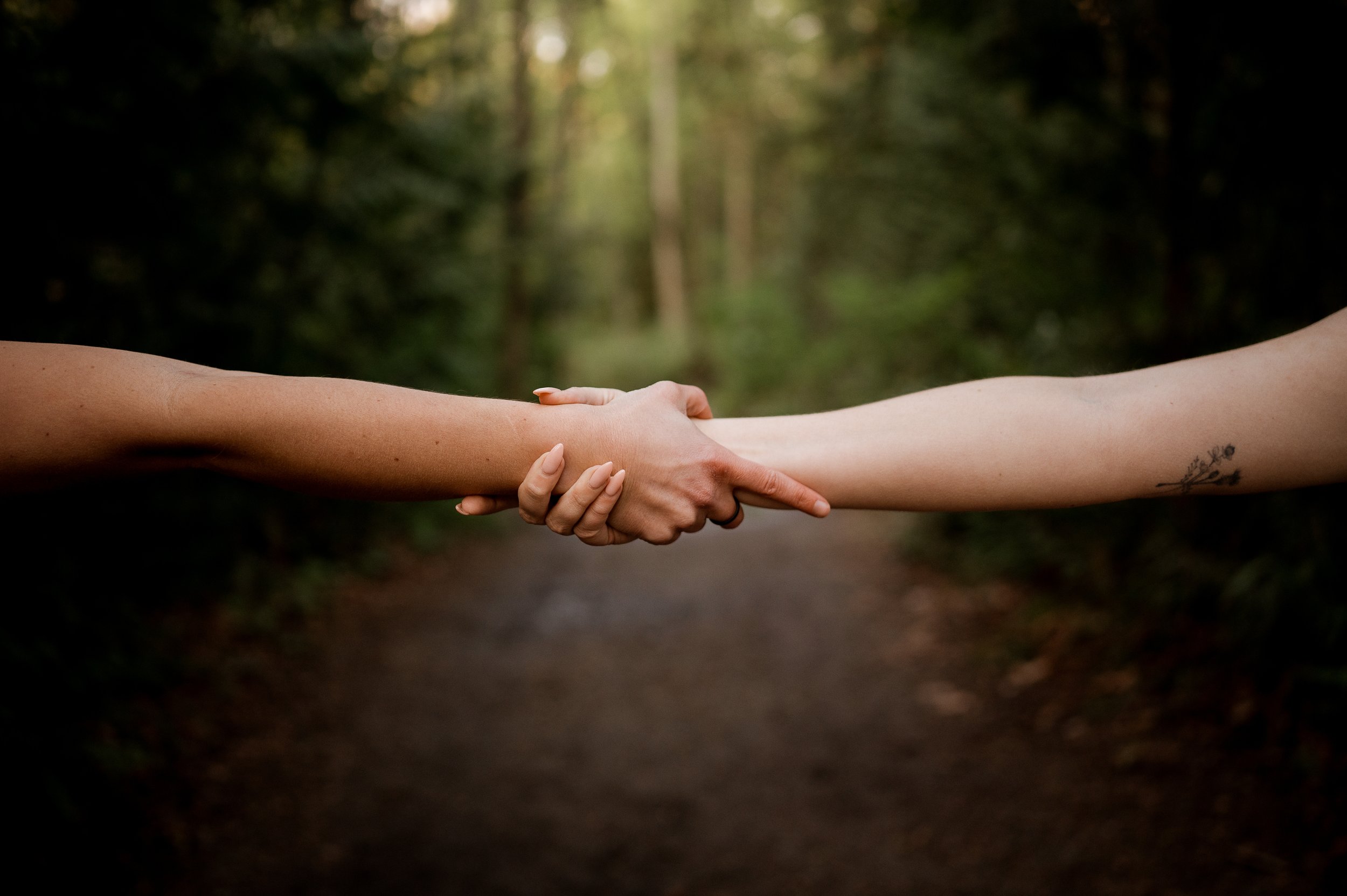 Two people holding hands with one person's arm tattooed, in a wooded outdoor setting.