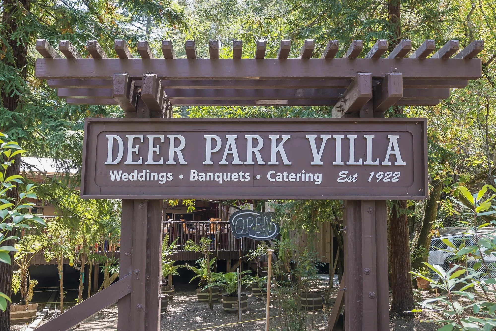 Wooden sign reading 'Deer Park Villa, Weddings, Banquets, Catering, Est 1922' with an 'Open' sign hanging below, surrounded by trees and greenery.