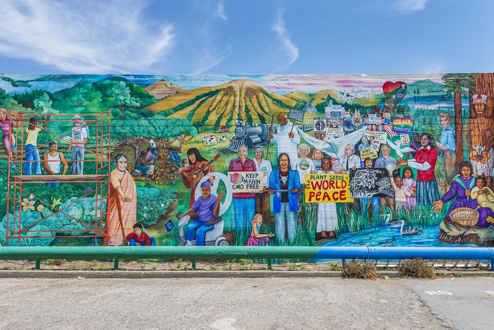 Colorful mural depicting diverse people protesting for peace, environmental protection, and social justice, with scenes of farming, activism, and community gathering against a backdrop of hills, mountains, and a clear blue sky.