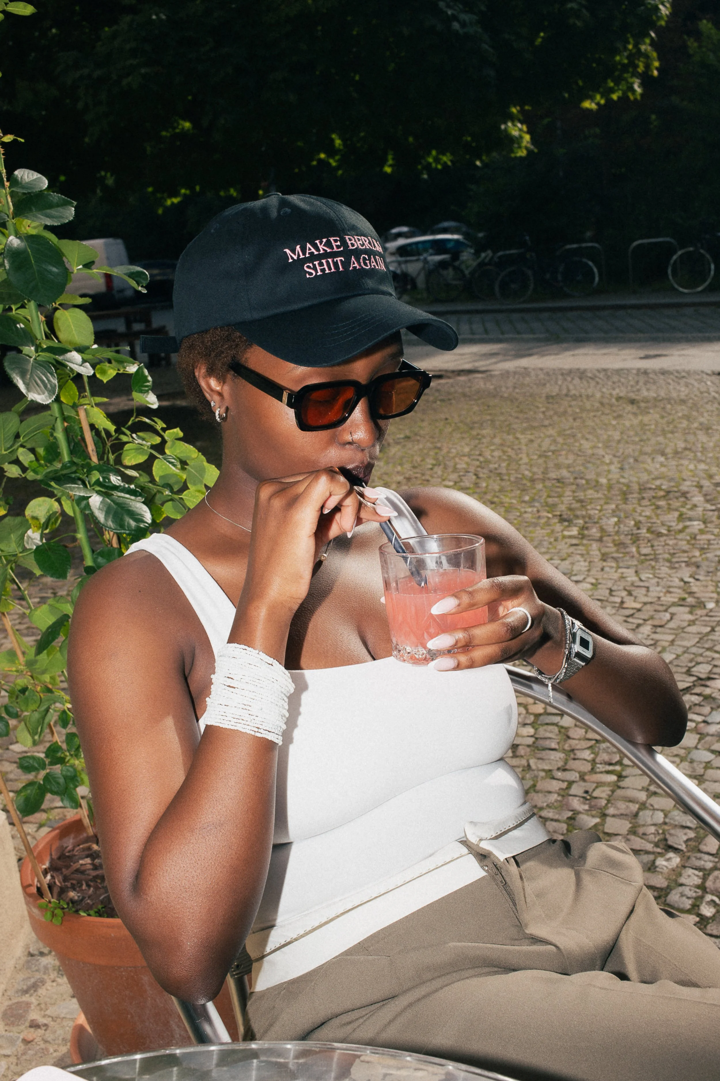 Berlin street style: A person wearing a statement black cap, sunglasses, and white tank top, sipping a matcha latte through a straw on a sunny outdoor terrace.