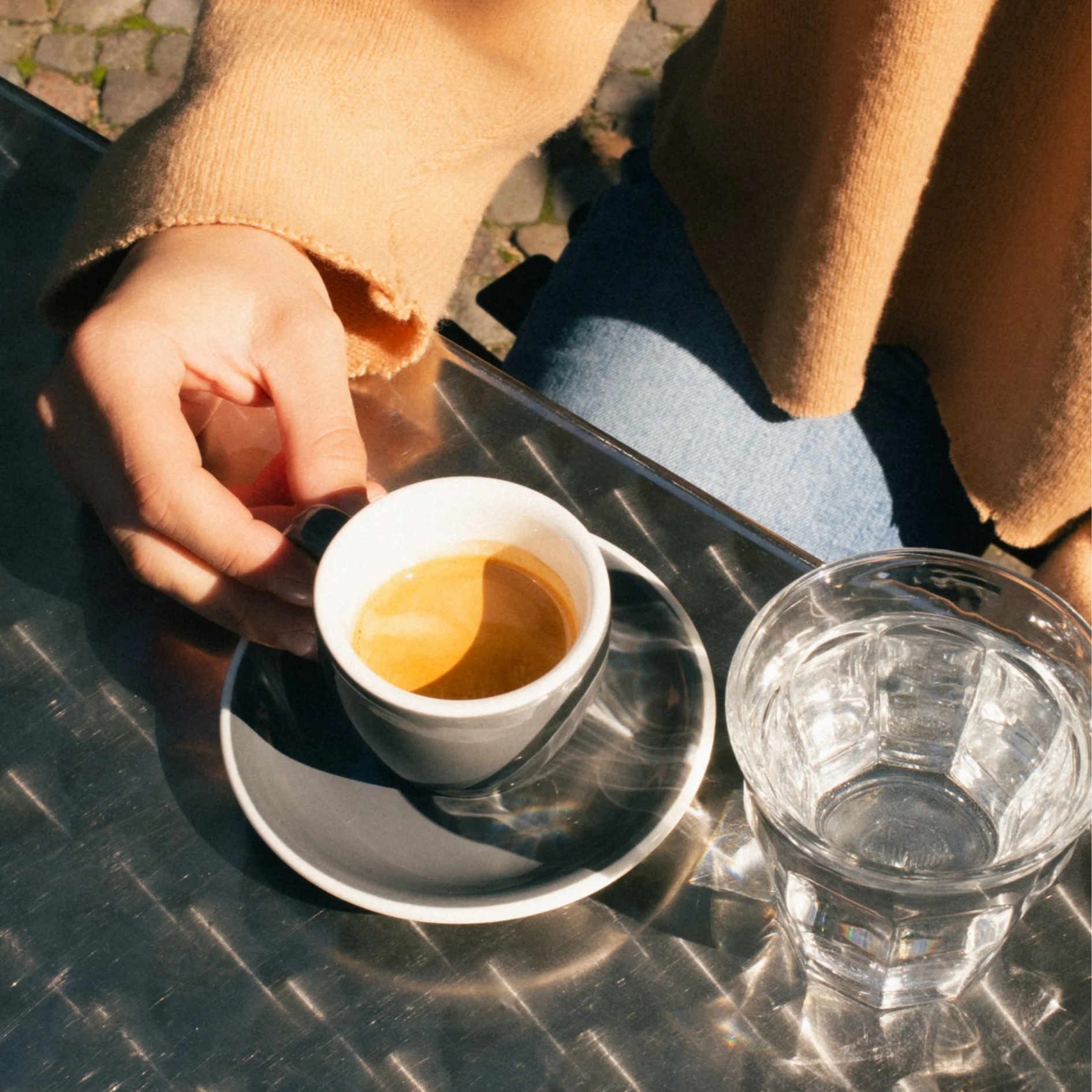 Person holding a small white espresso cup with a dark saucer, next to a empty glass of water, on a shiny metal table outside.