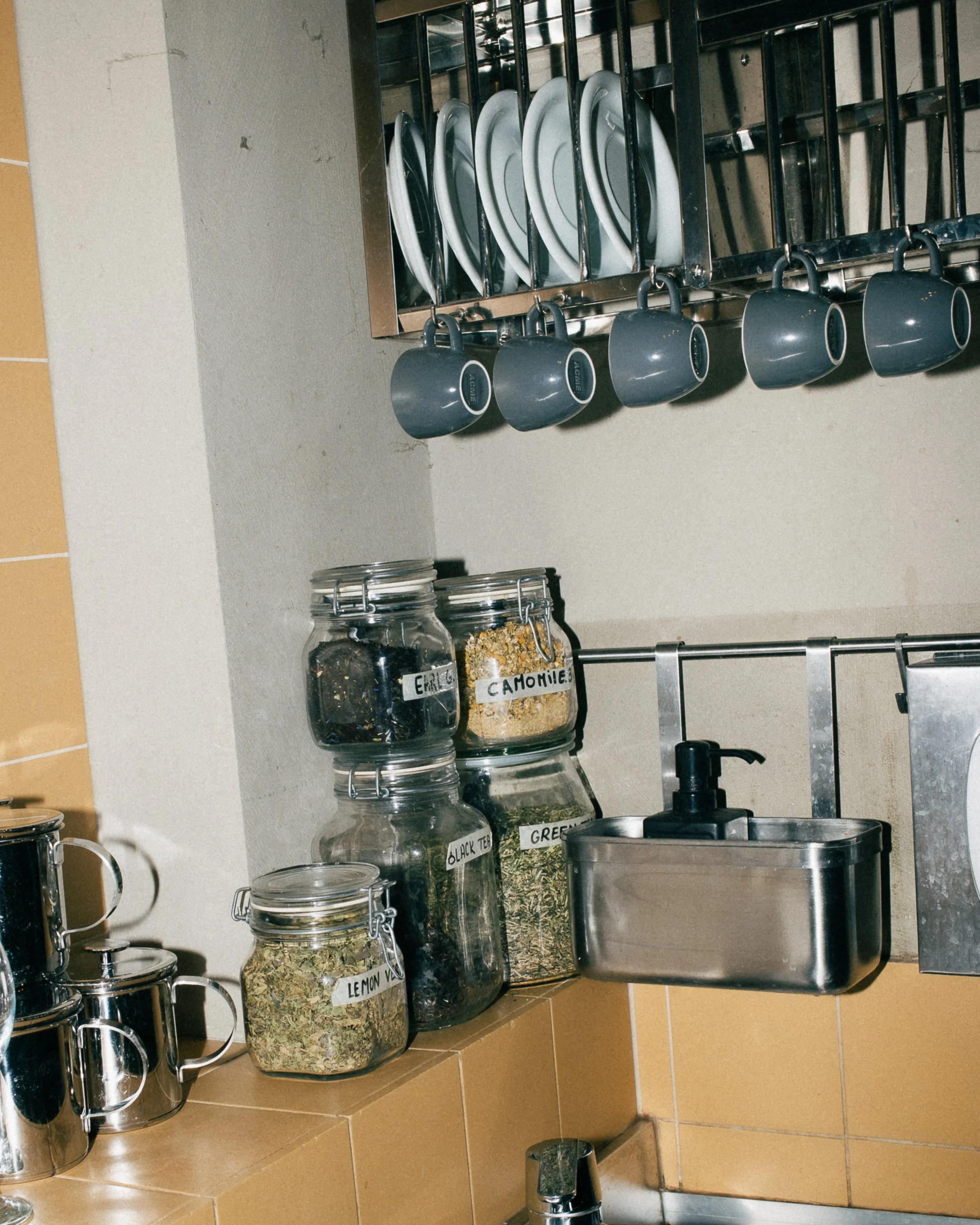 Minimalist kitchen setting in a specialty coffee shop with a stainless steel sink, hanging mugs, and an aesthetic display of tea ingredients in glass jars.