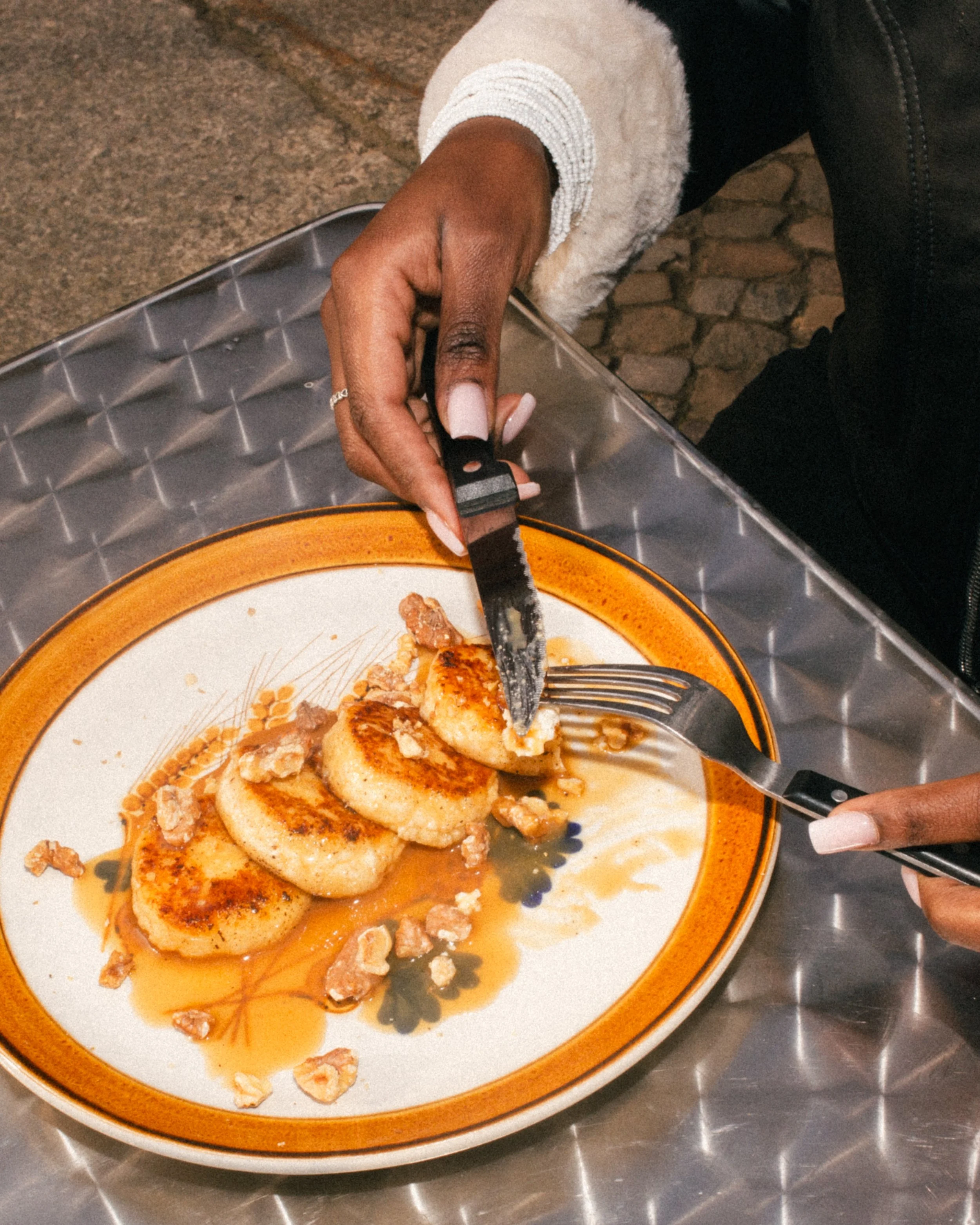 Person cutting into three toasted cheese pancakes topped with honey and sprinkled with nuts on a decorative plate, sitting on a metal table.