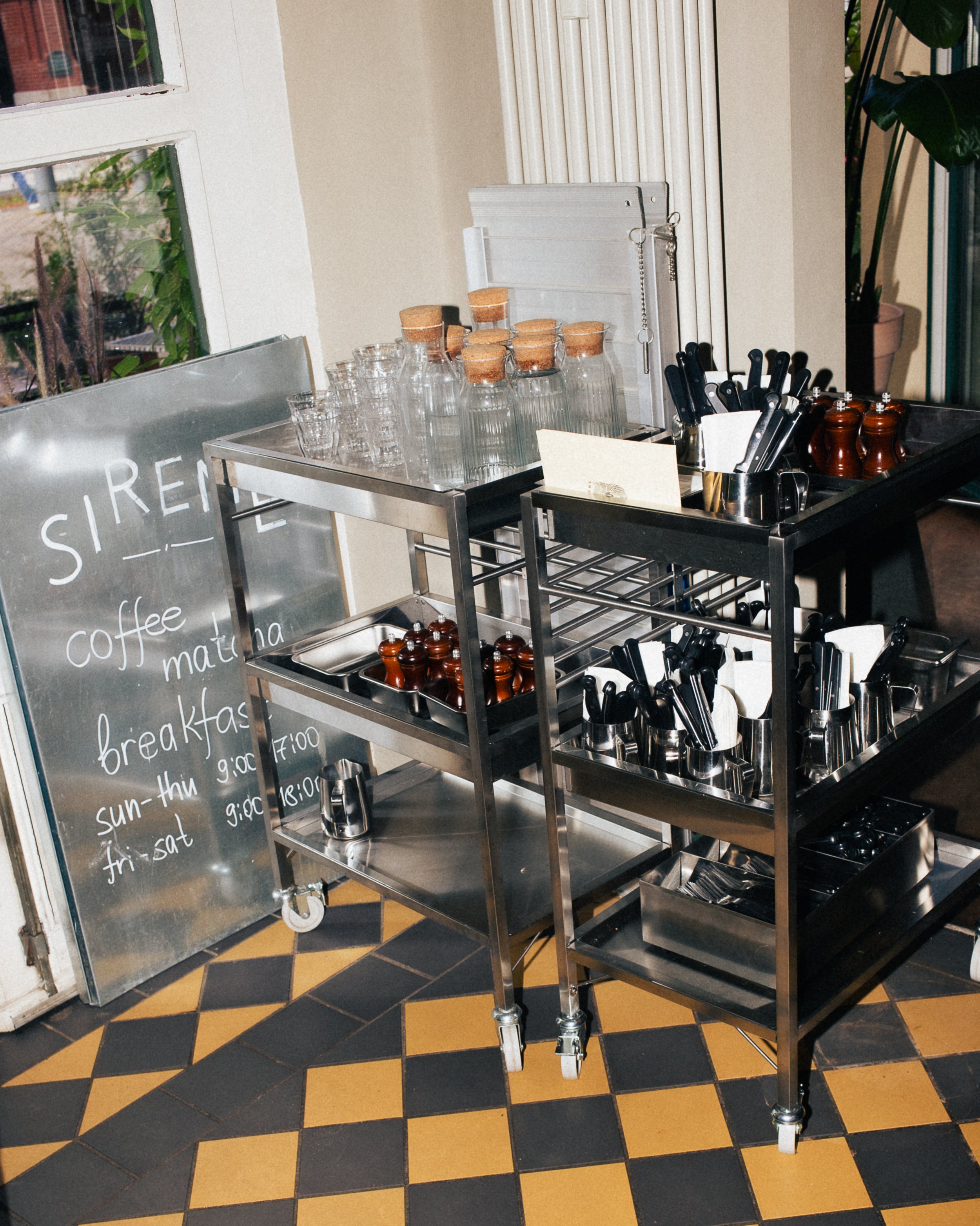 A stainless steel catering cart with various jars, cups, utensils, and condiments, next to a small chalkboard menu listing coffee and breakfast hours.