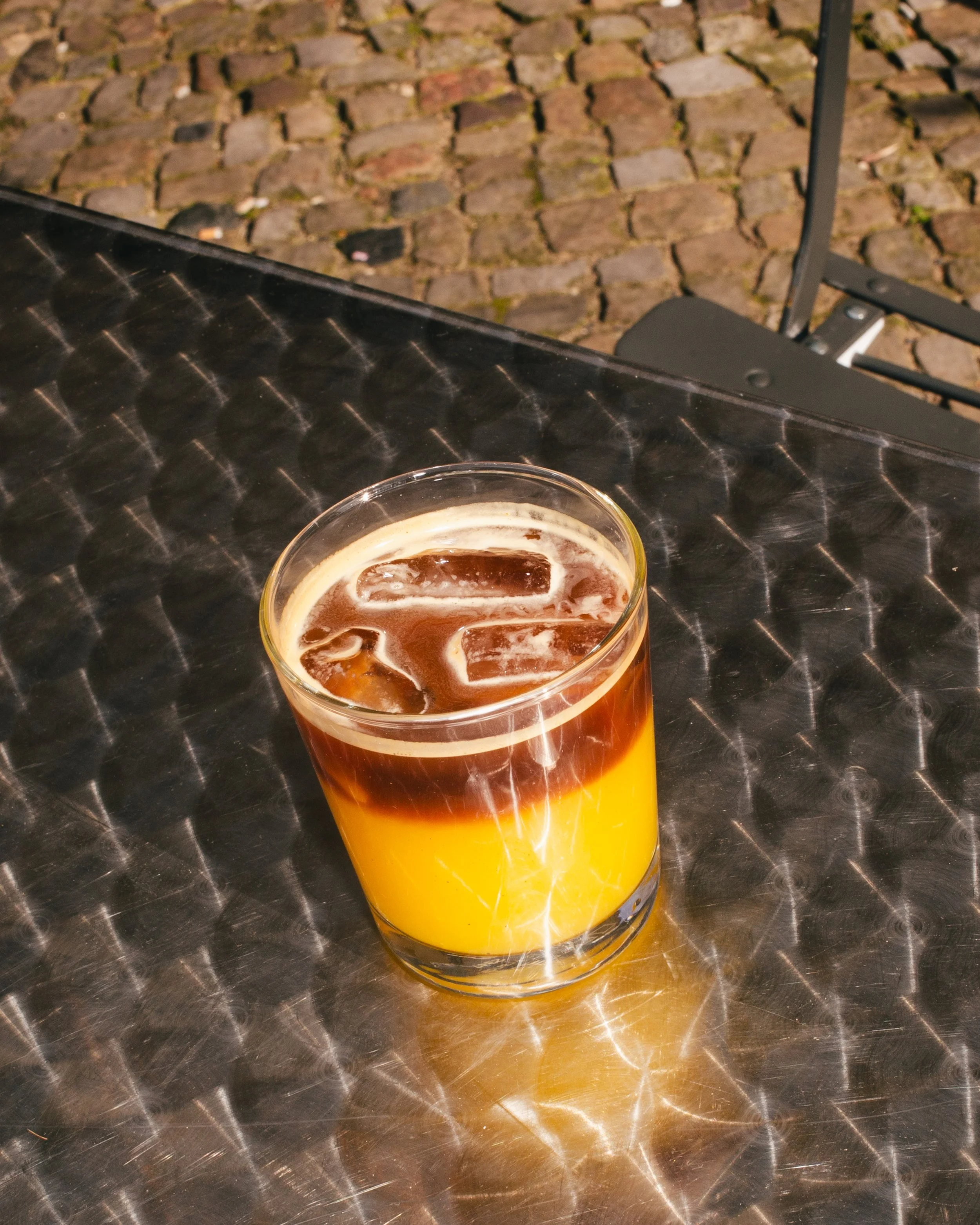 A glass of a layered drink with a yellow orange juice layer and a espresso top layer, containing ice cubes, on a textured metal table outside on a cobblestone surface.