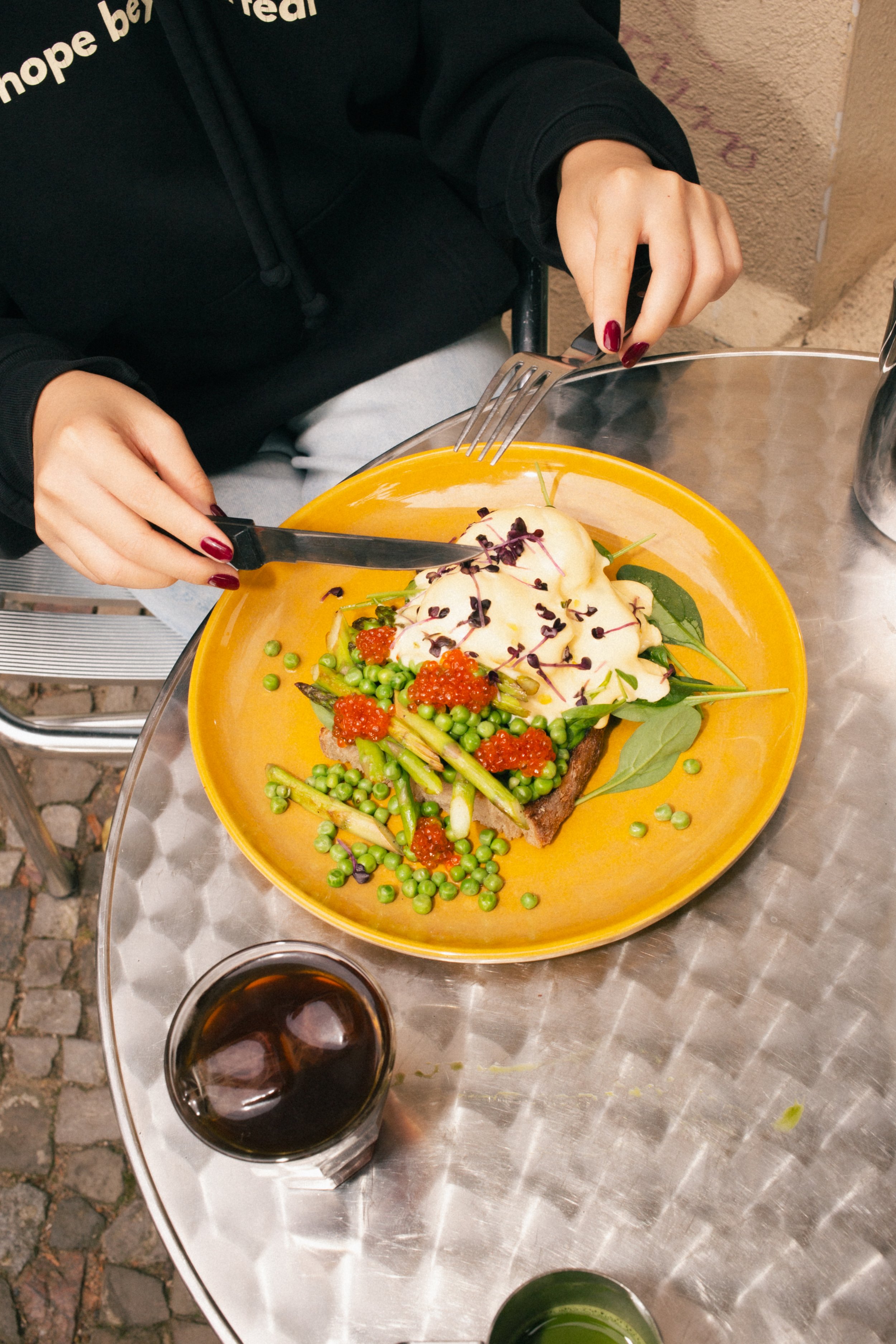 A person in a black hoodie with red painted nails is cutting into a plated meal of toast with seasonal asparagus and eggs with a creamy topping and red caviar on a yellow plate, on a silver outdoor table with a glass of dark soda.