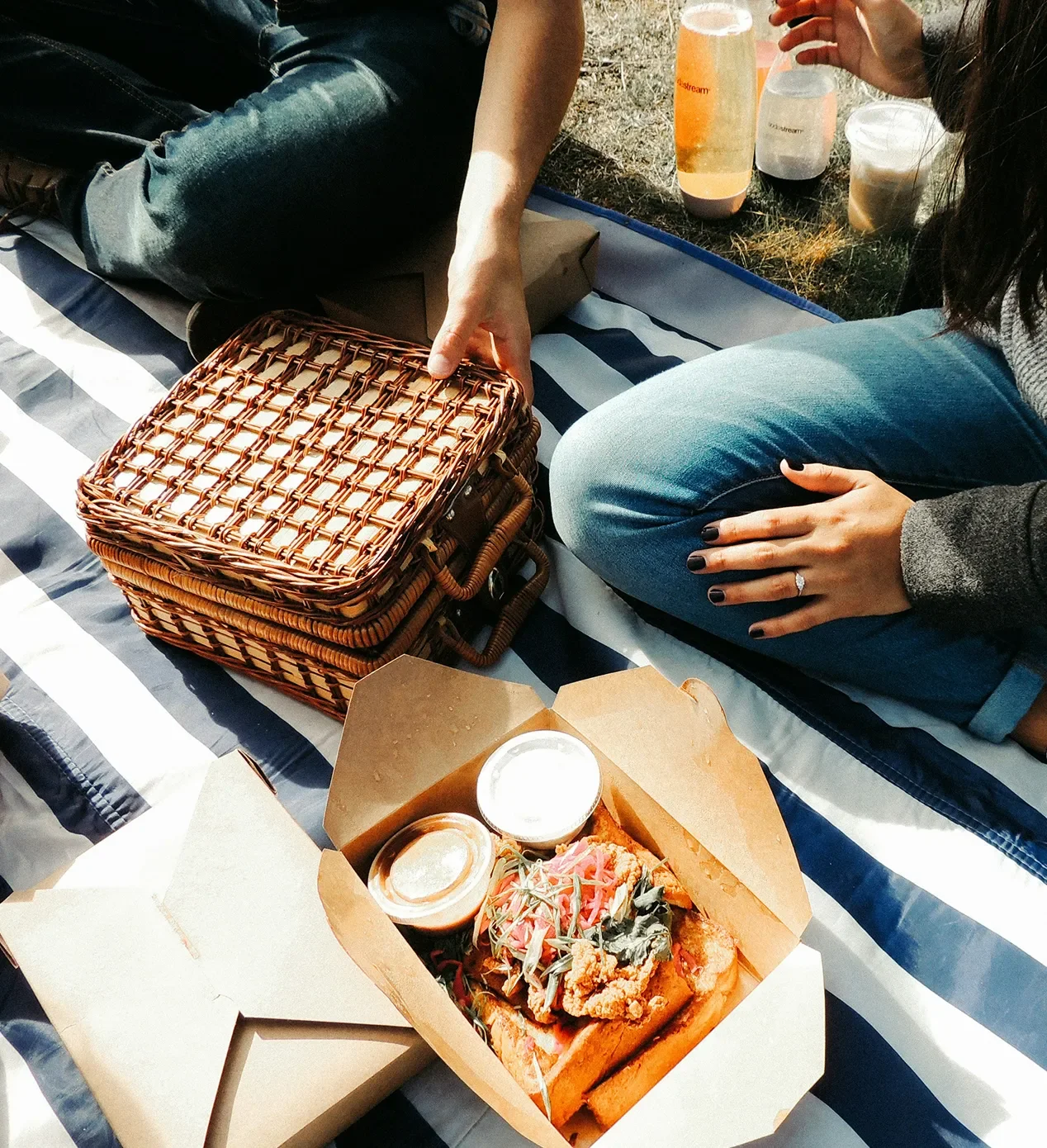 People enjoying a picnic outdoors with a wicker basket, a box of food including fried chicken and sauces, and drinks on a blanket.