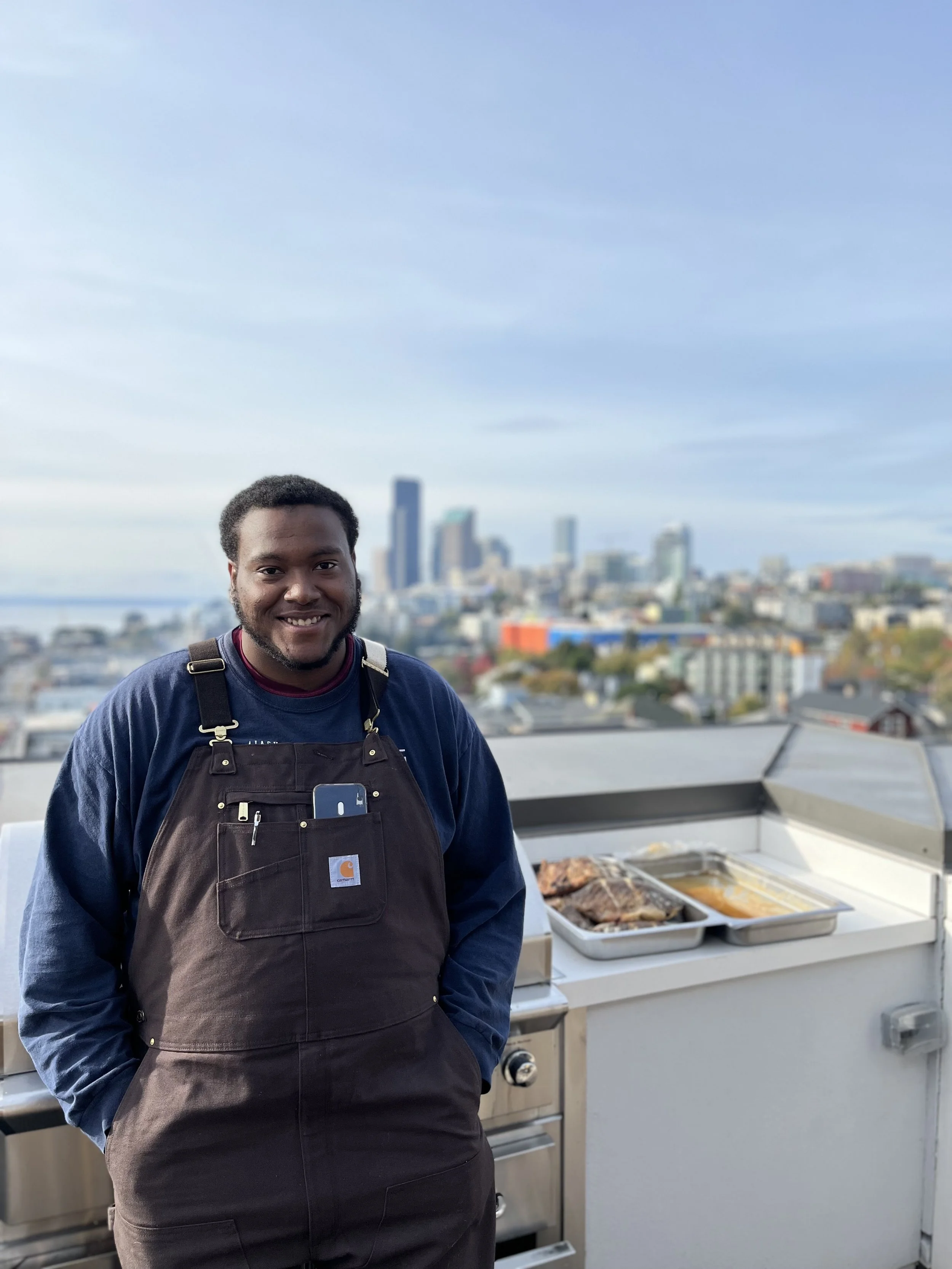 A man standing in front of a rooftop barbecue grill with city skyline in the background.