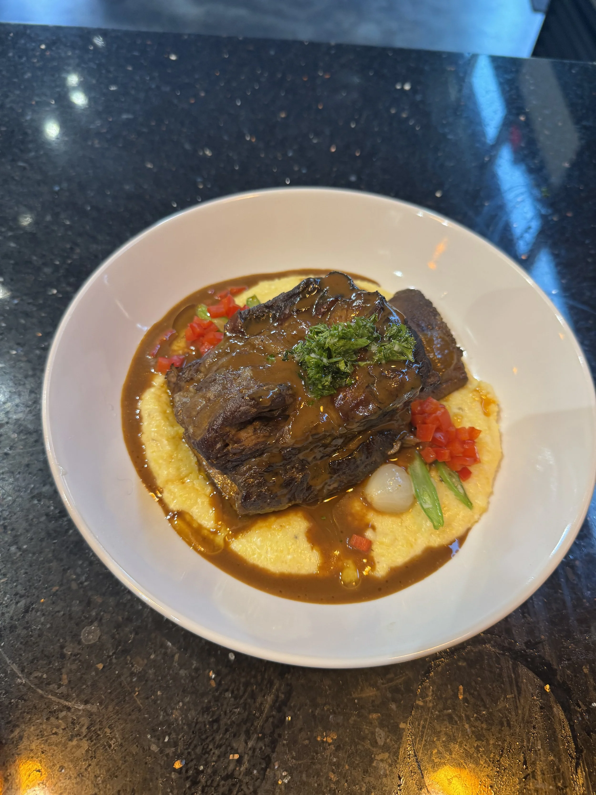 Plate of braised beef ribs served over creamy mashed potatoes, garnished with chopped parsley and surrounded by diced red peppers and green onions.