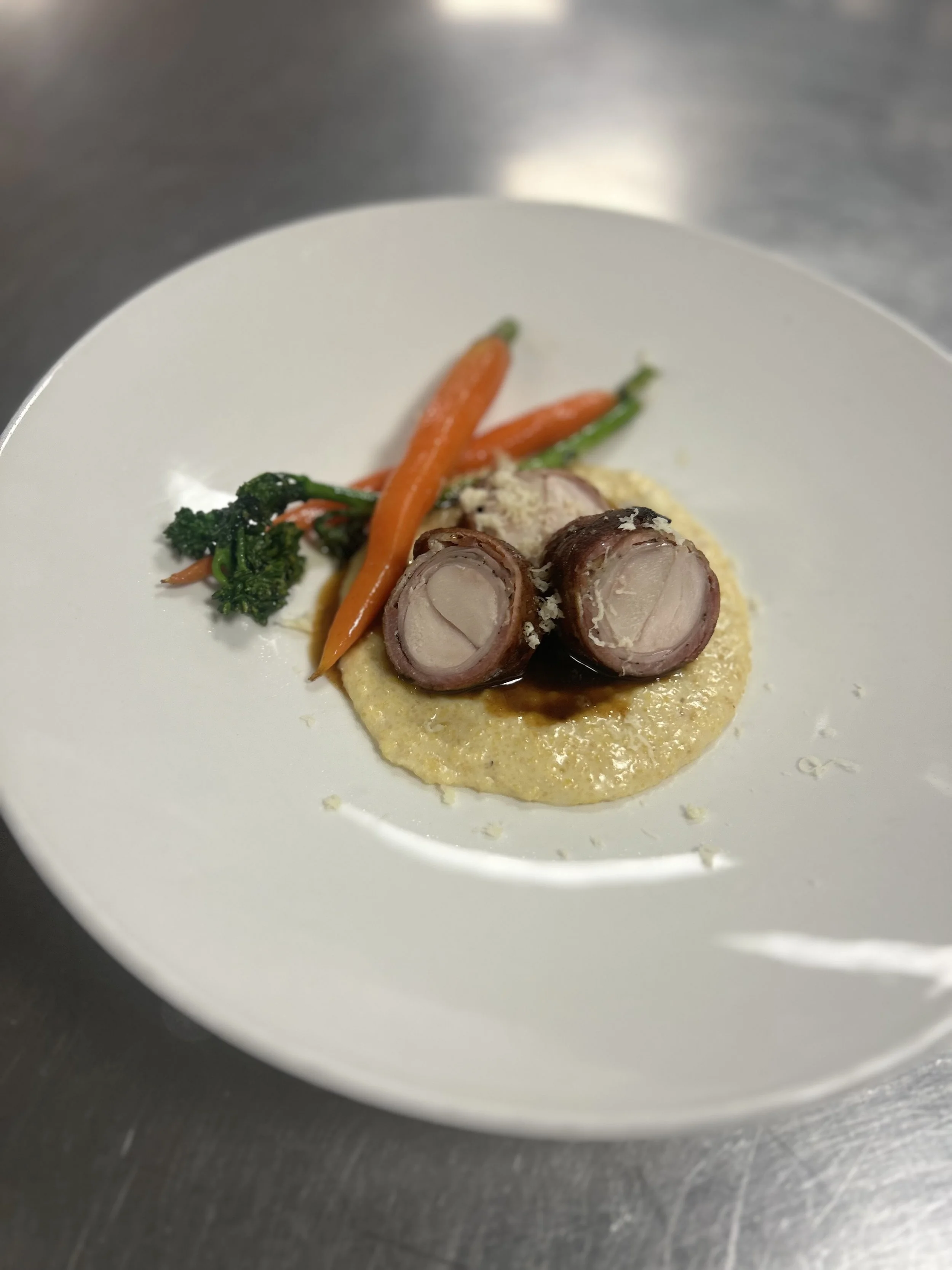 A plated gourmet dish featuring sliced rolled meat, creamy puree, and sautéed carrots and broccoli on a white plate.