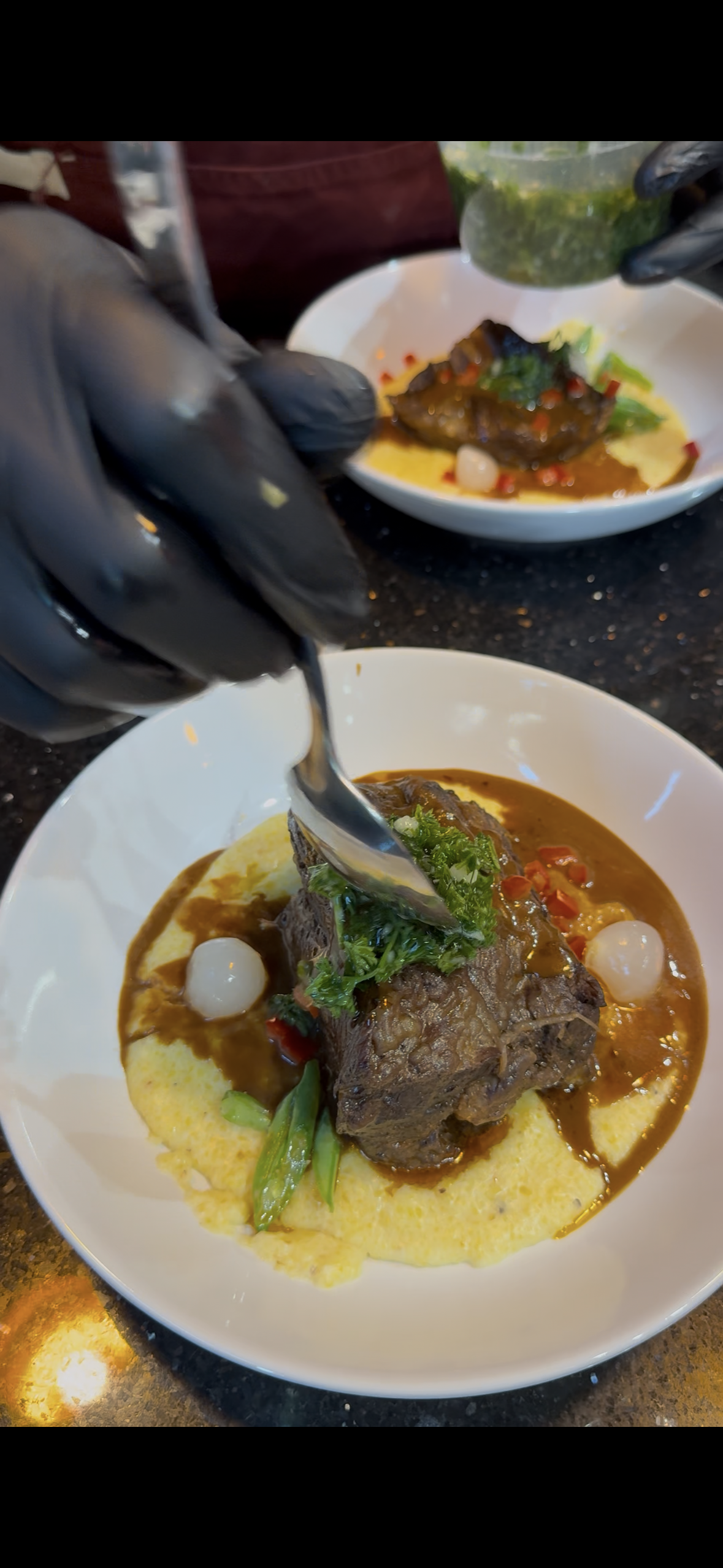 A plated dish featuring a large piece of braised meat on a bed of creamy mashed potatoes, garnished with cilantro, green onions, and a dark sauce.