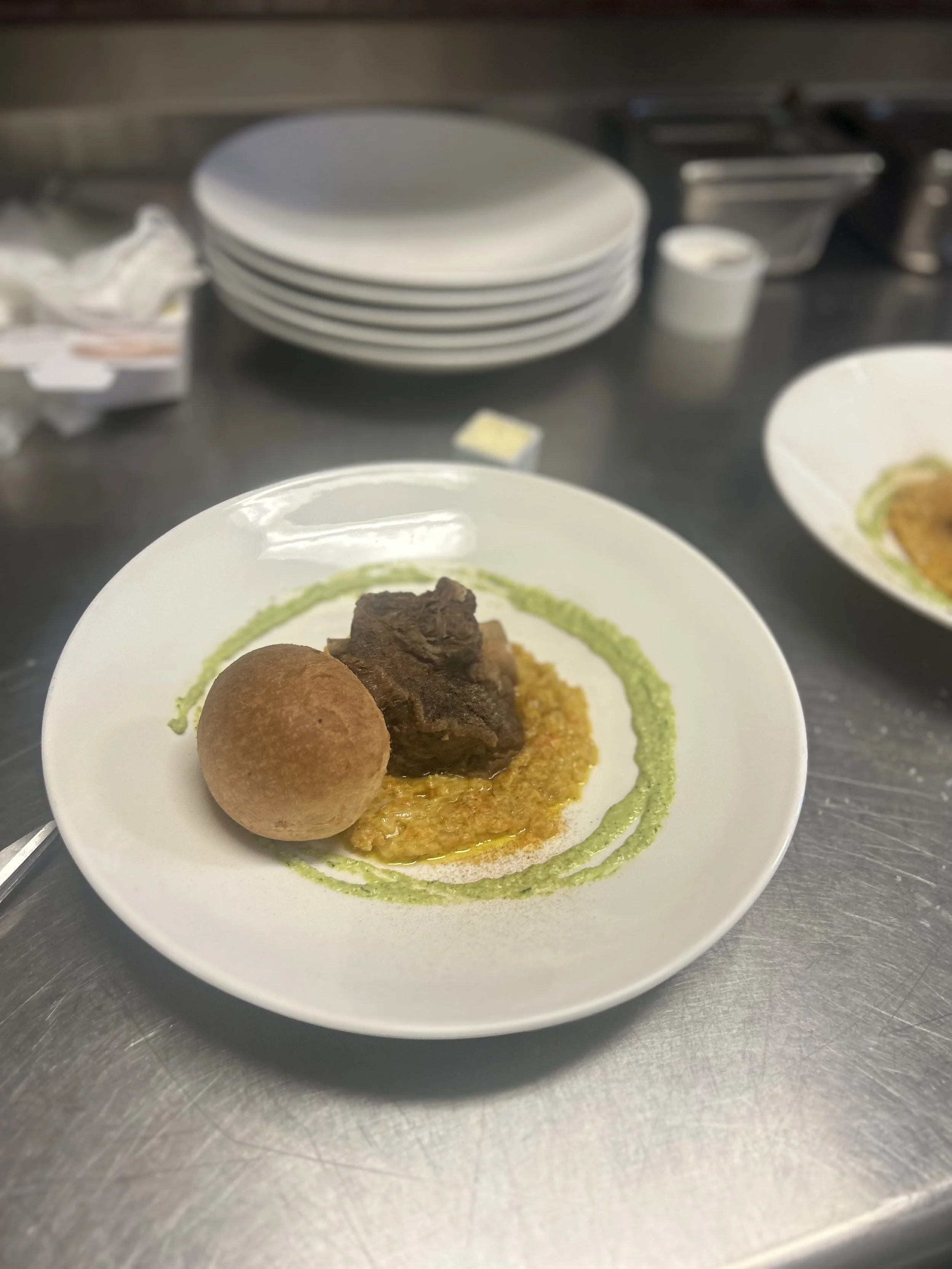 A white plate with a meat dish, a potato ball, and a bed of yellow sauce, garnished with green sauce in a swirl pattern on a stainless steel countertop.