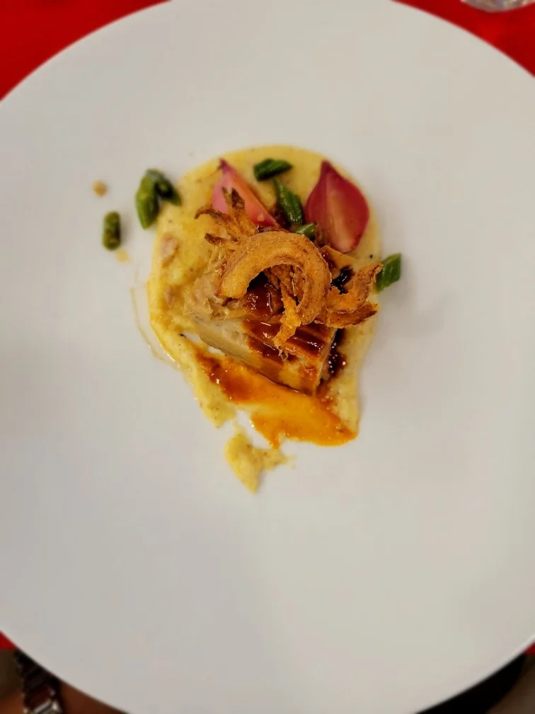 A plated gourmet dish featuring a portion of fish topped with fried onions, served on a bed of creamy mashed potatoes with a drizzle of sauce, garnished with tomato slices and green onions.