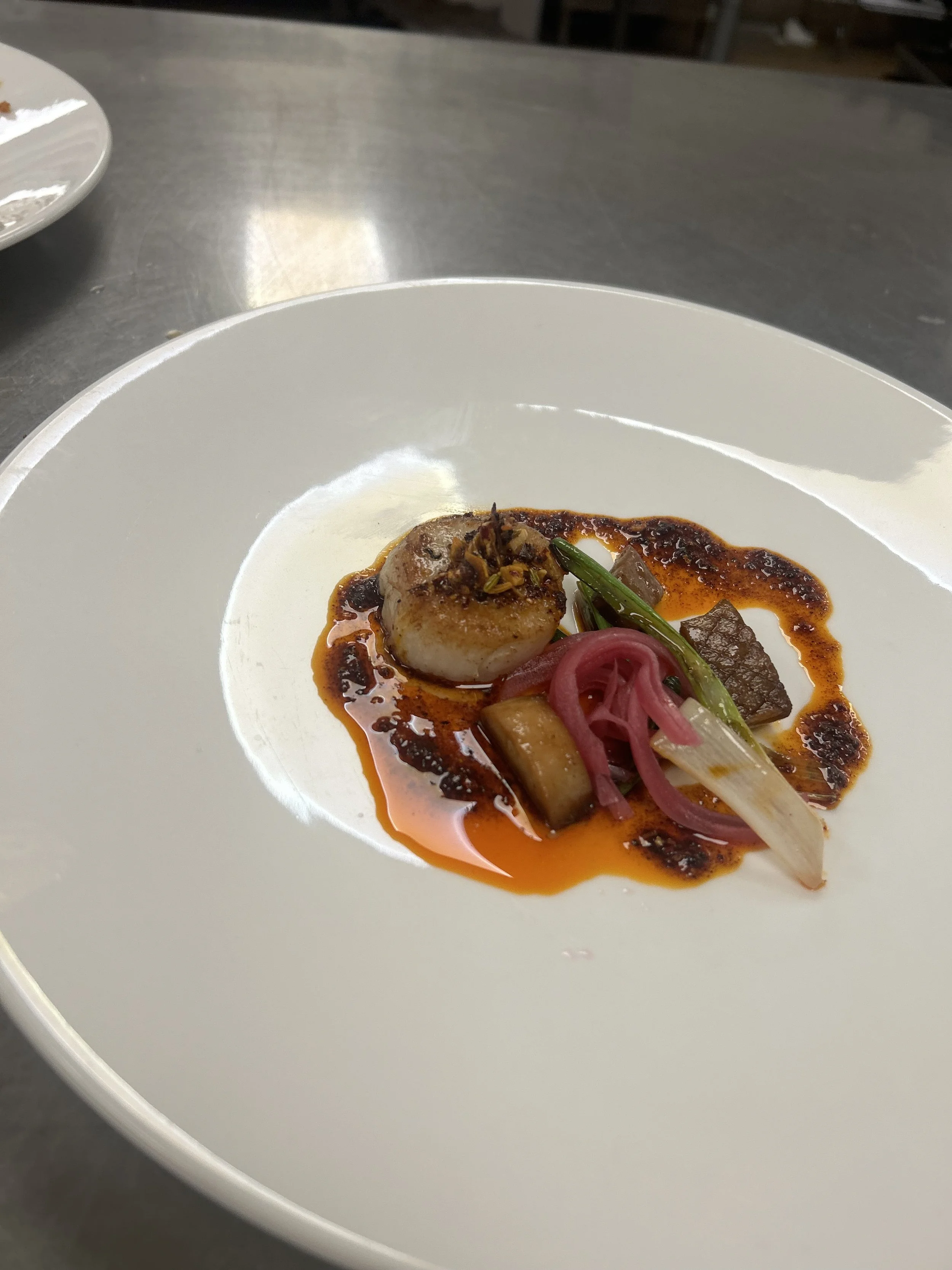 A plated dish with a seared scallop topped with herbs, accompanied by roasted vegetables including eggplant, green beans, and pickled red onions, all in a reddish sauce.