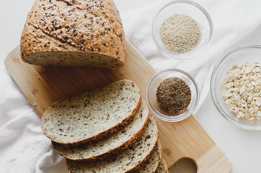 Sliced multigrain bread on a wooden cutting board with bowls of quinoa, seeds, and oats placed beside it.