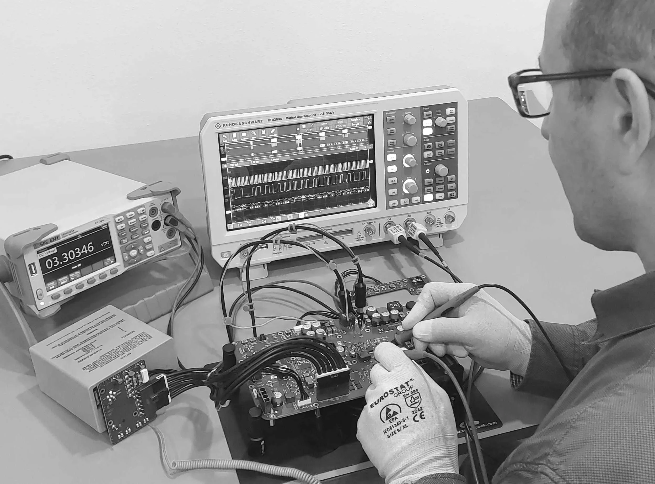 Hardware design workspace with oscilloscope and custom PCB prototypes. Engineer measures on the board with probes.